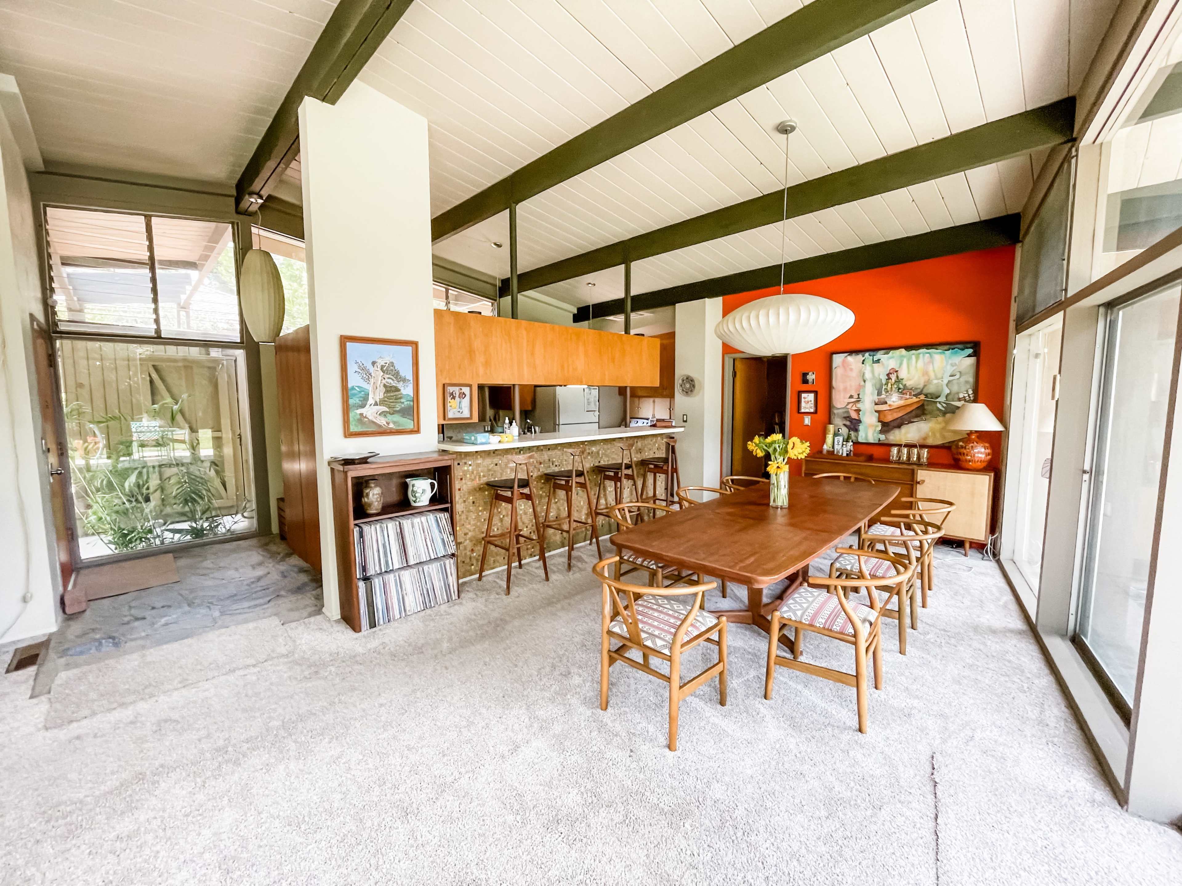 The image shows a mid-century modern dining area with a long wooden table, rattan chairs, and a vibrant accent wall.