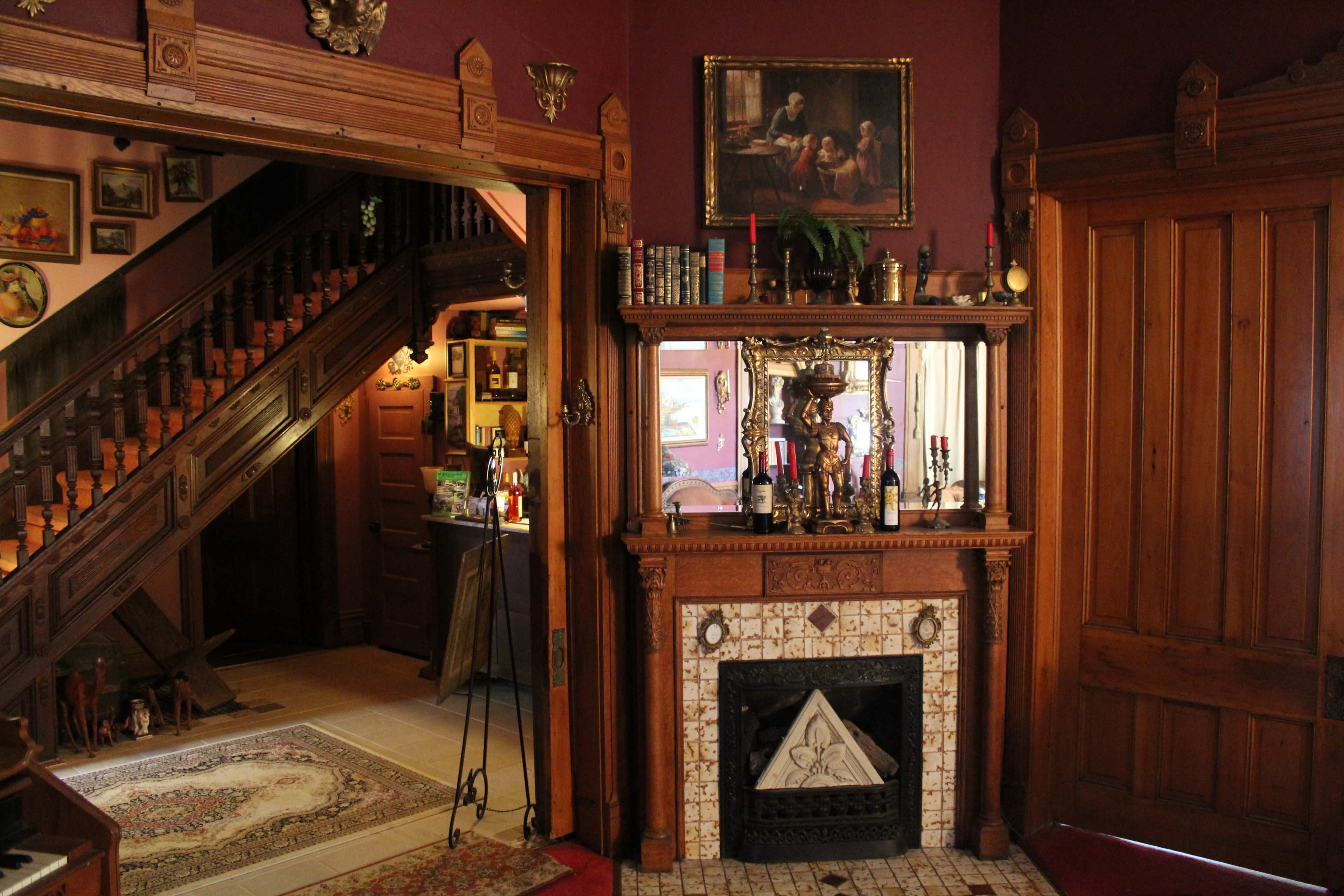 The image shows a richly decorated interior featuring a wooden staircase, a fireplace with ornate decorations, and walls adorned with artwork and shelves.