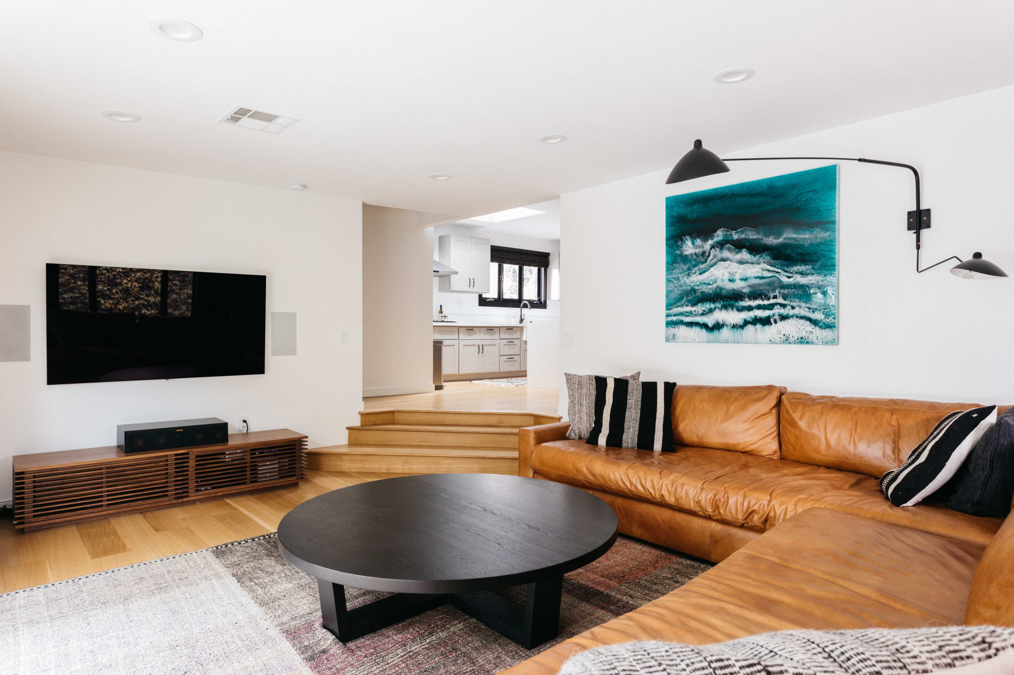 A modern living room features a brown leather sectional sofa, a circular black coffee table, and a large wall-mounted television, with a kitchen visible in the background.