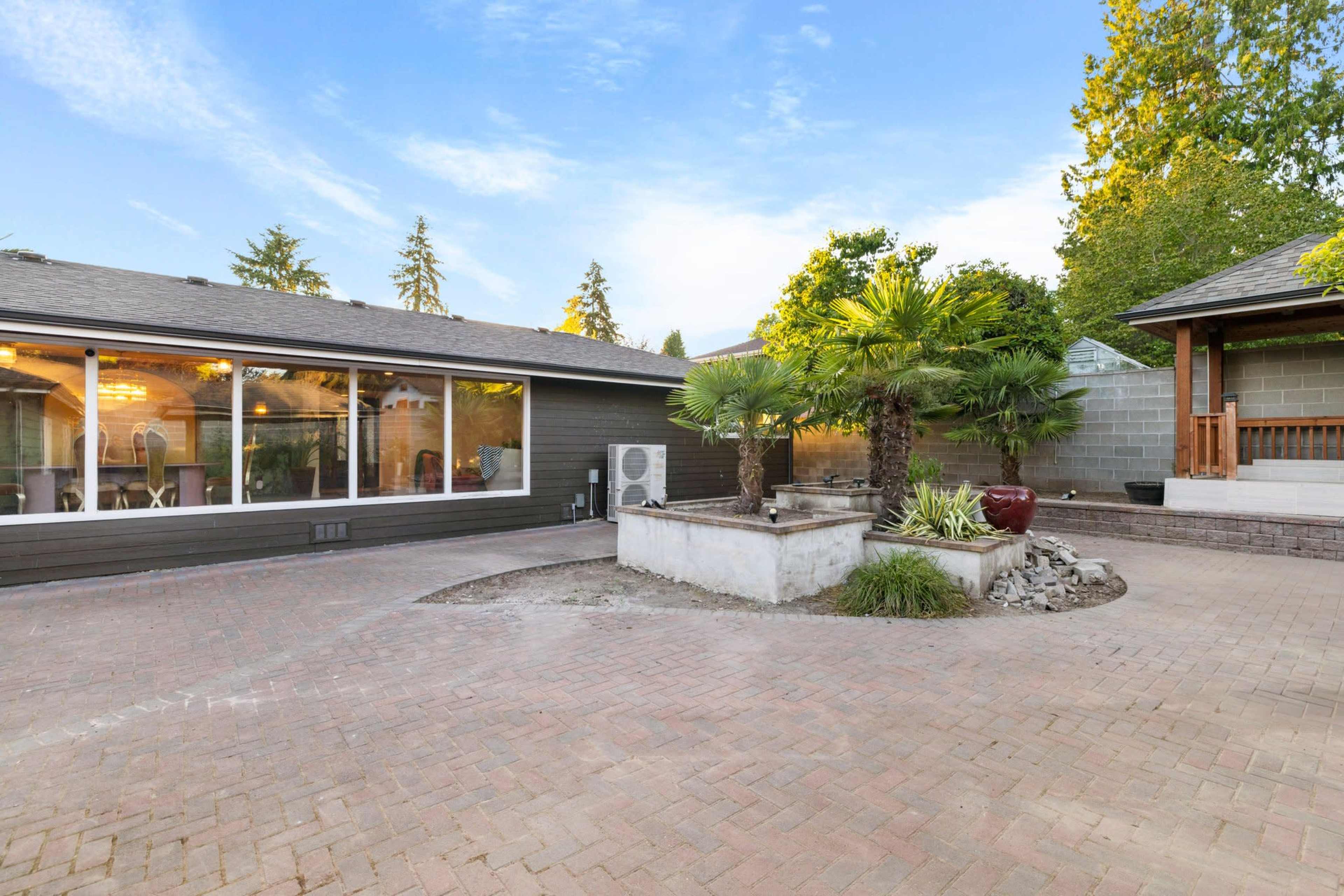 A spacious outdoor patio area featuring a brick surface, a small fountain with plants, and a neighboring structure with a shaded porch.