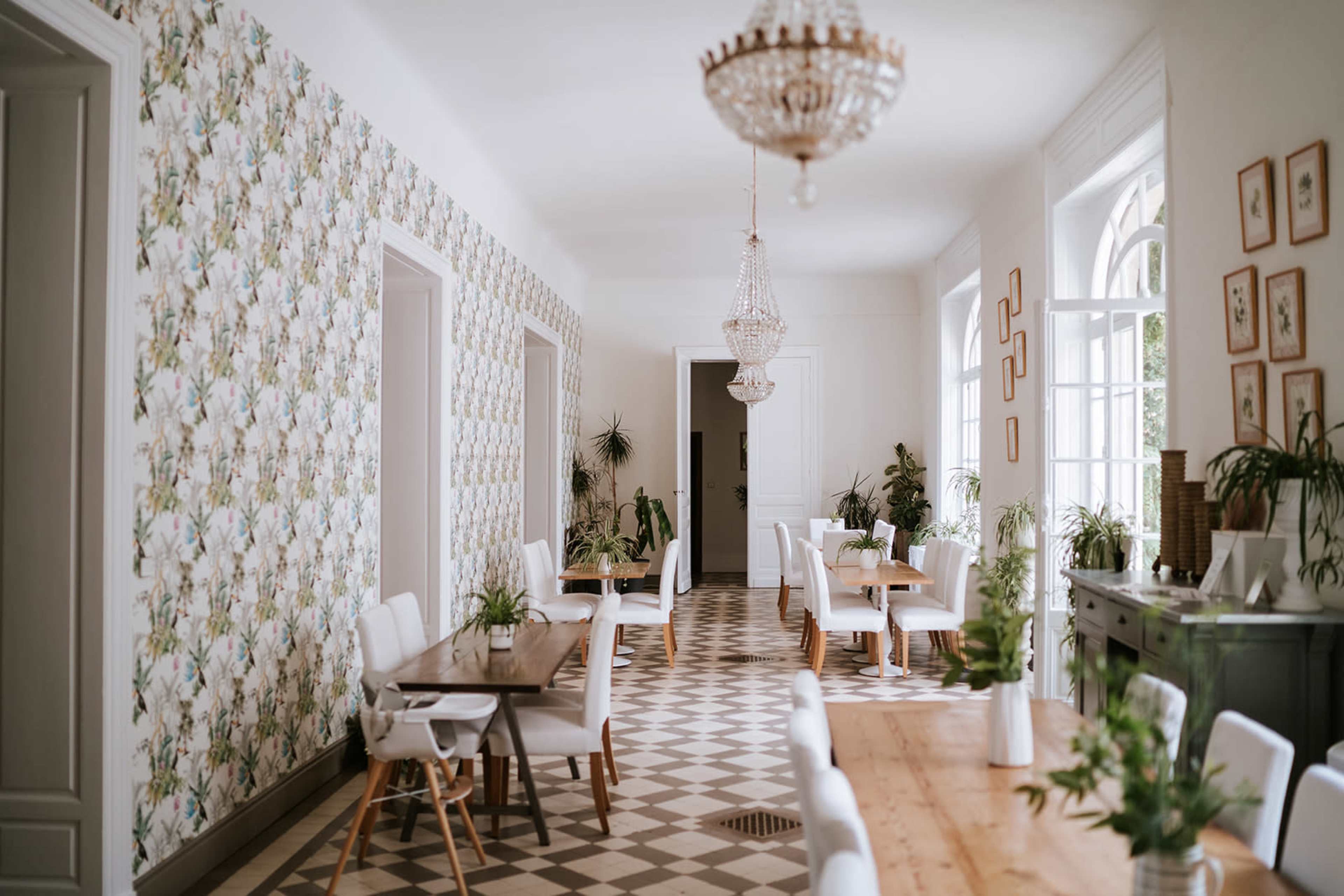The image shows a well-lit dining area with patterned wallpaper, rectangular wooden tables, and white chairs arranged neatly along the sides.