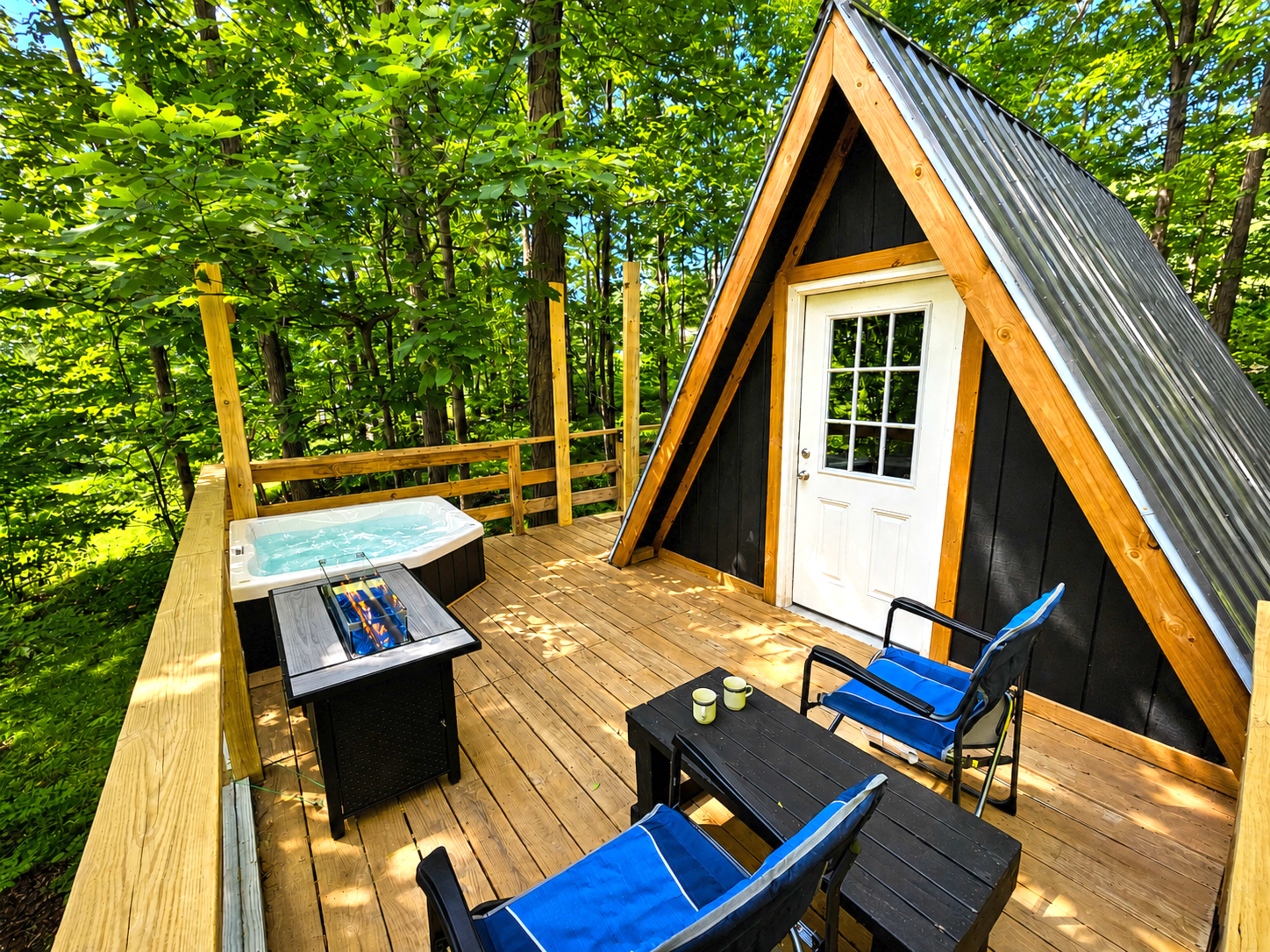 A modern A-frame cabin with a hot tub and seating area is set on a wooden deck surrounded by trees.