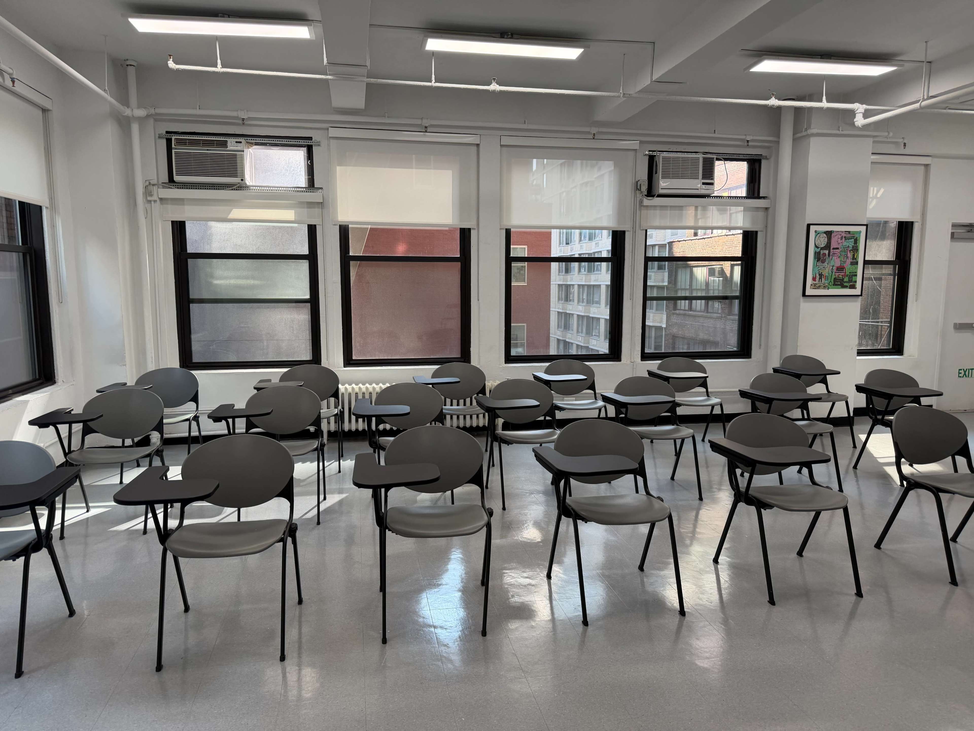 The image shows a classroom with rows of black chairs arranged around a bright, empty space with large windows.