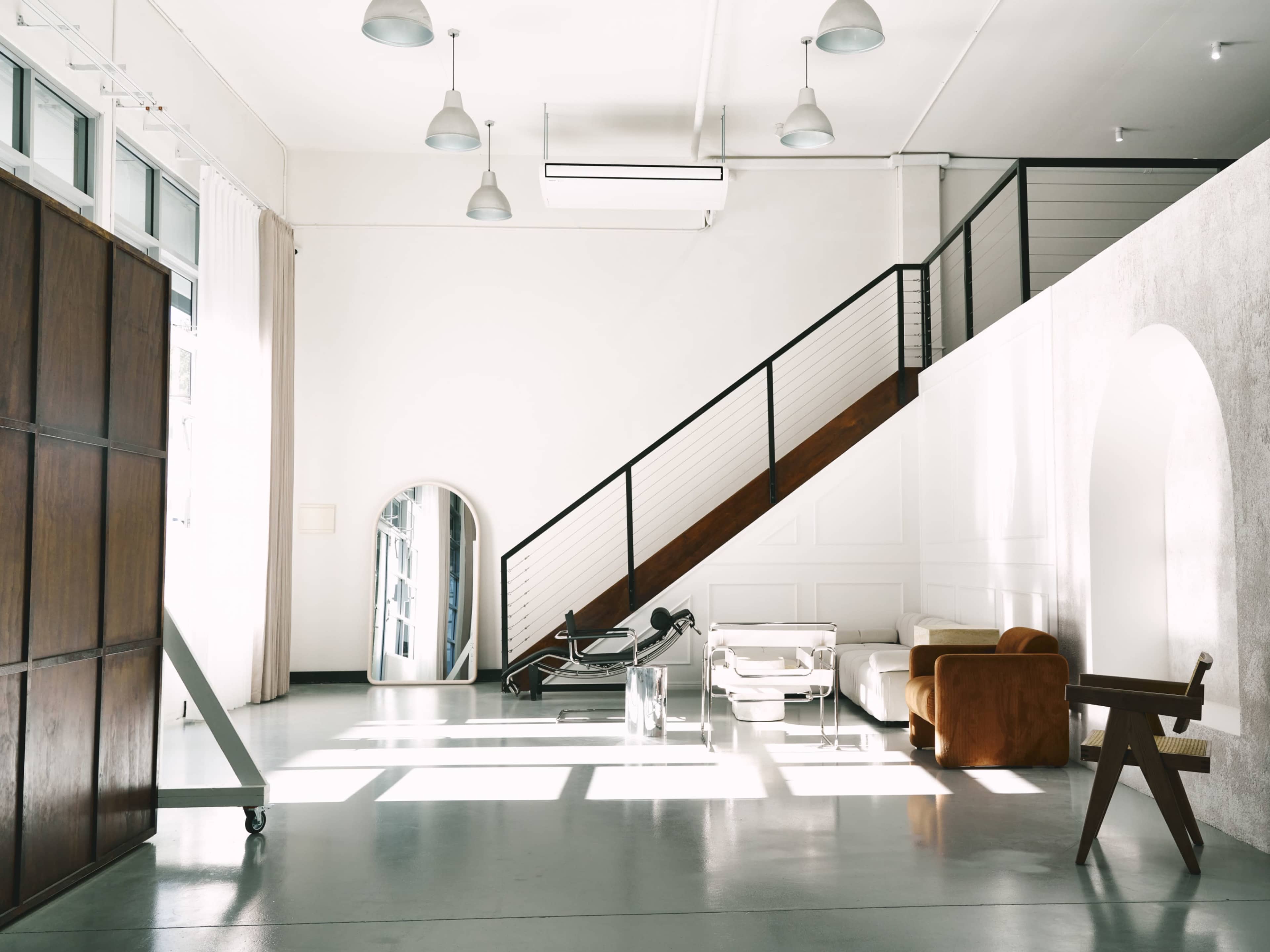 The image depicts a spacious, modern interior featuring a staircase, minimalist furniture, and large windows allowing natural light to enter.