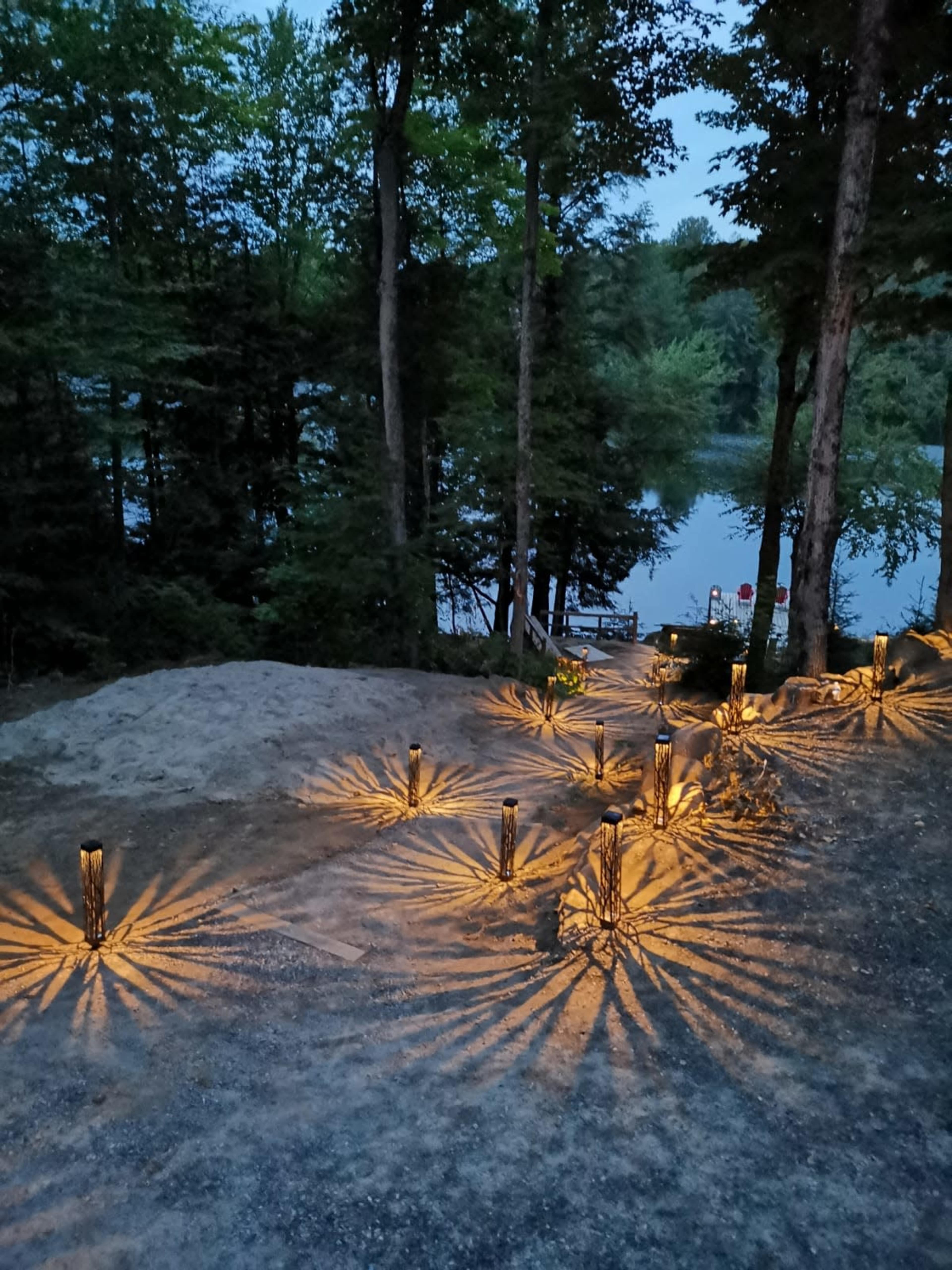 A wooded path leading to a lake, illuminated by tiki-style lights casting patterned shadows on the ground.