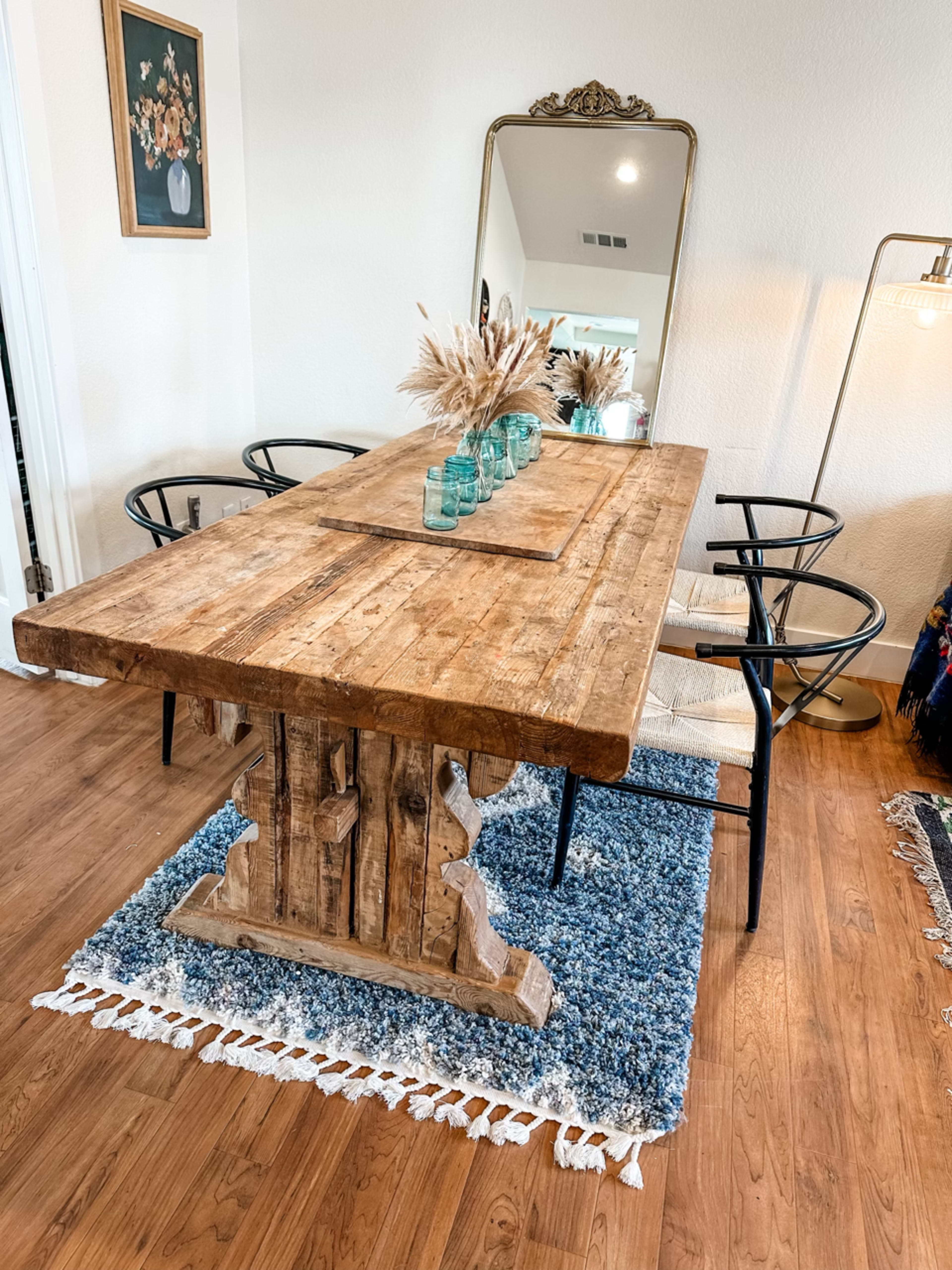 A rustic wooden dining table is set in a well-lit room with a large mirror, surrounded by black chairs and a decorative rug.