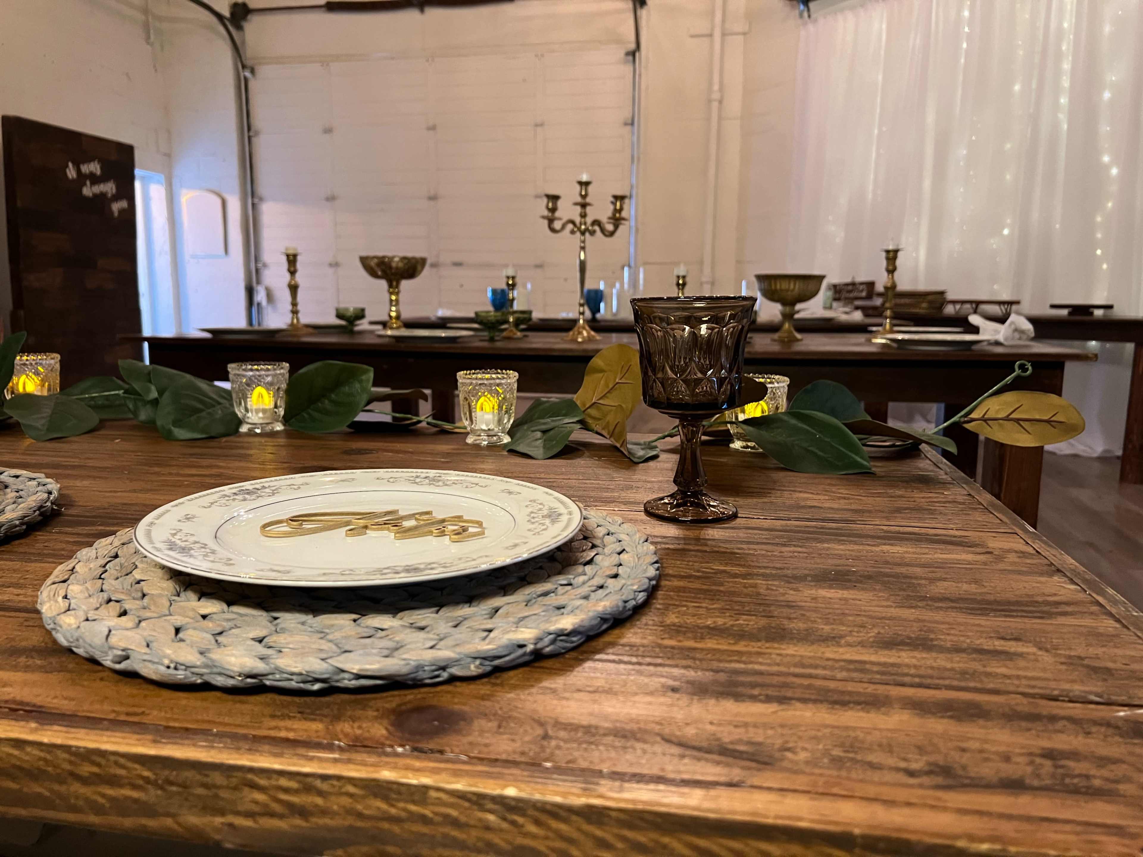 A dining setup with woven placemats, patterned plates, decorative glassware, and candles on a wooden table in a spacious room.
