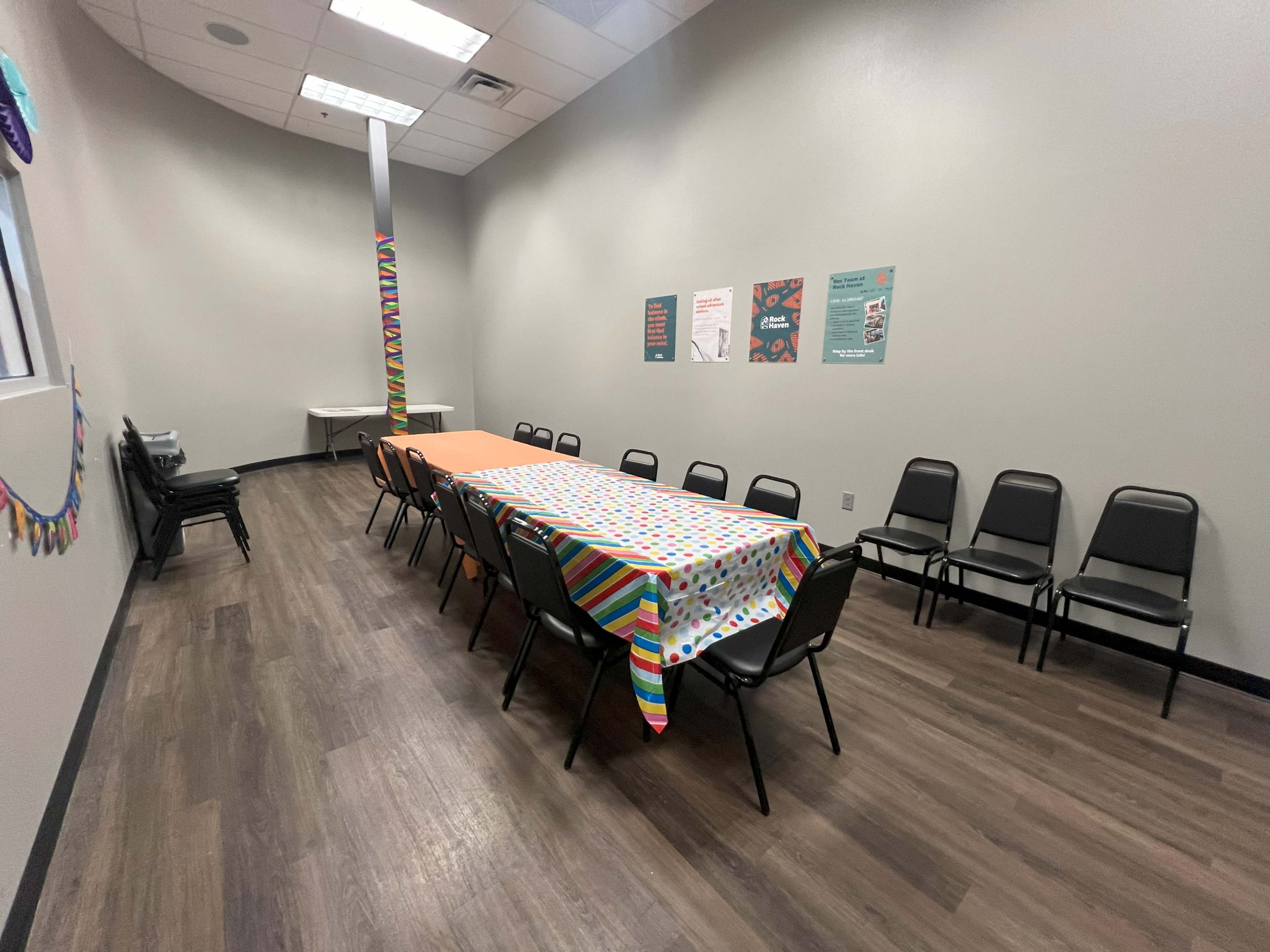 A long table covered with a patterned tablecloth is set up in a room with gray walls and black chairs arranged around it.