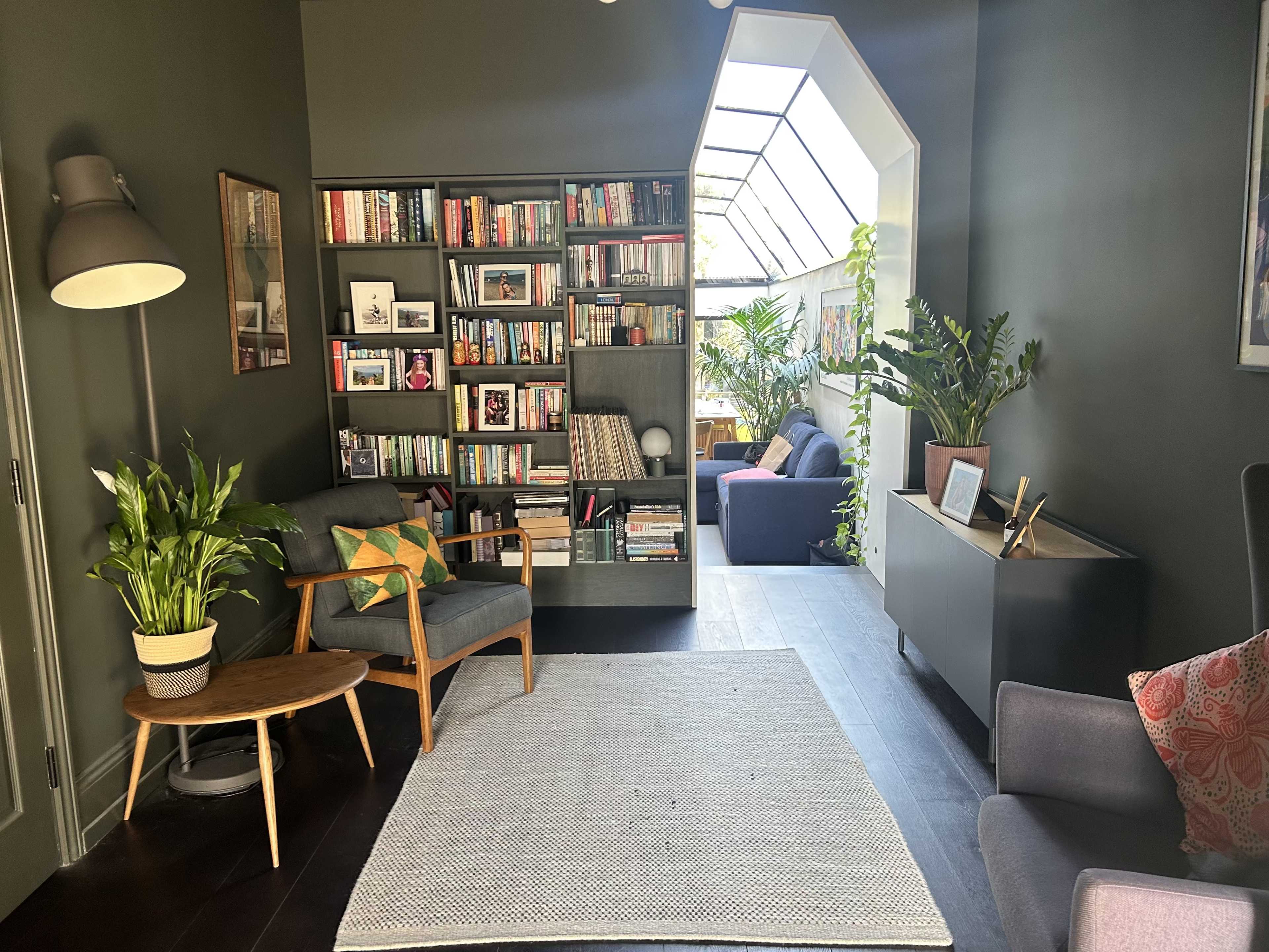 The image shows a cozy living space featuring a bookshelf, indoor plants, and comfortable seating arranged around a light-filled area with a glass roof.