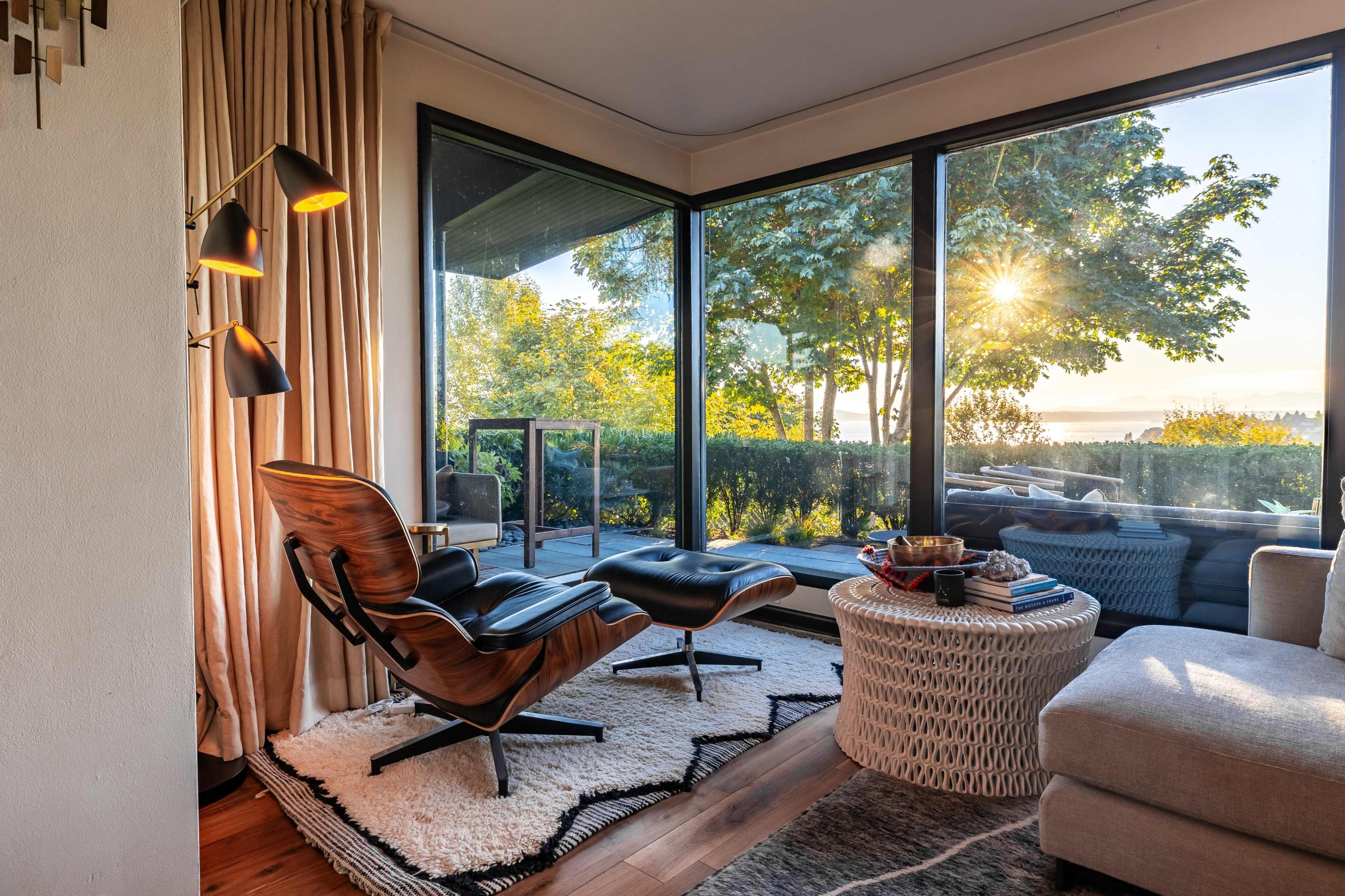 A cozy living room features two chairs beside a round table, with large windows offering a view of a tree-lined outdoor space and a sunset.