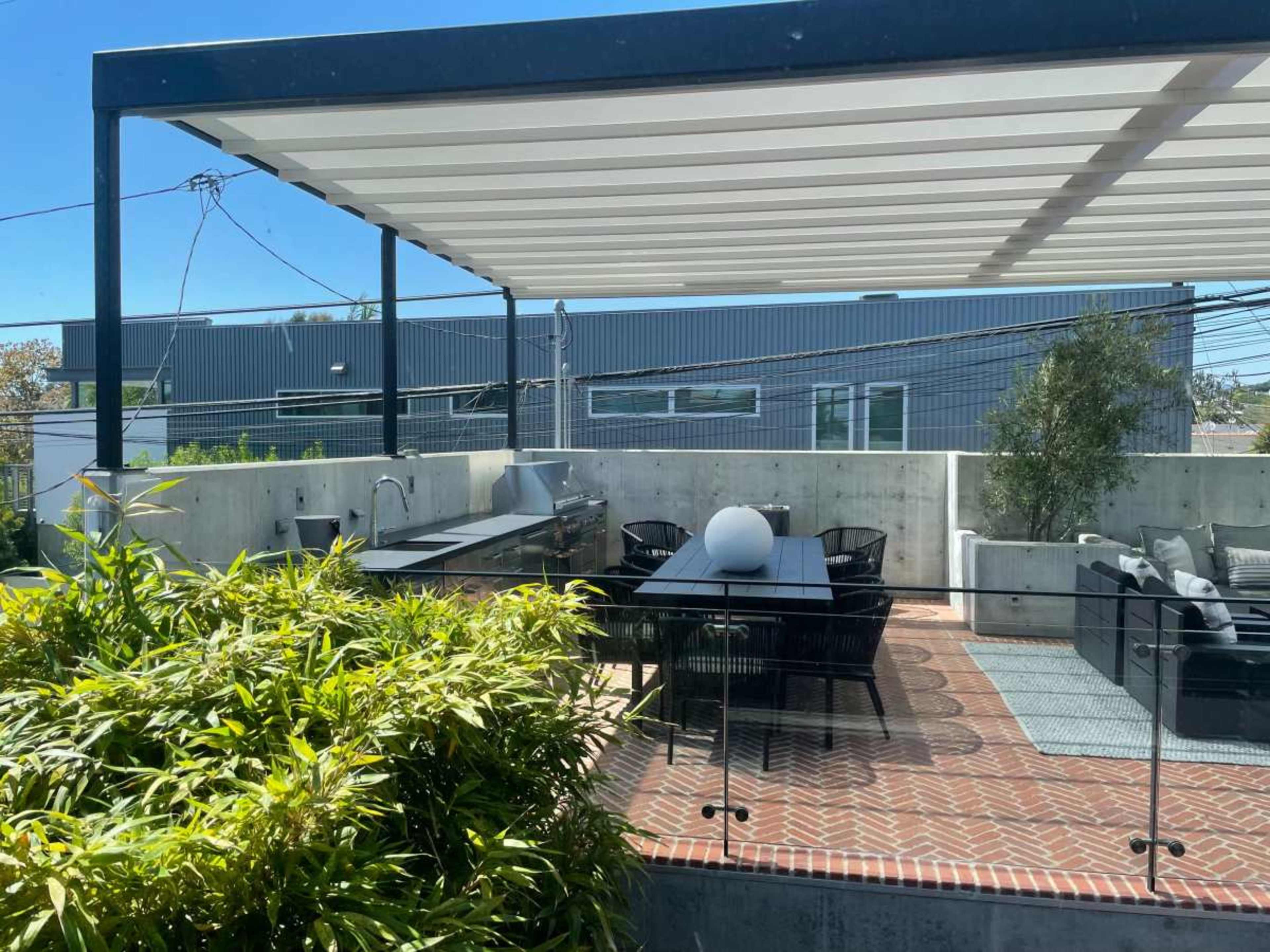 A modern outdoor patio area with a dining table, seating, and a built-in grill, surrounded by concrete walls and greenery.