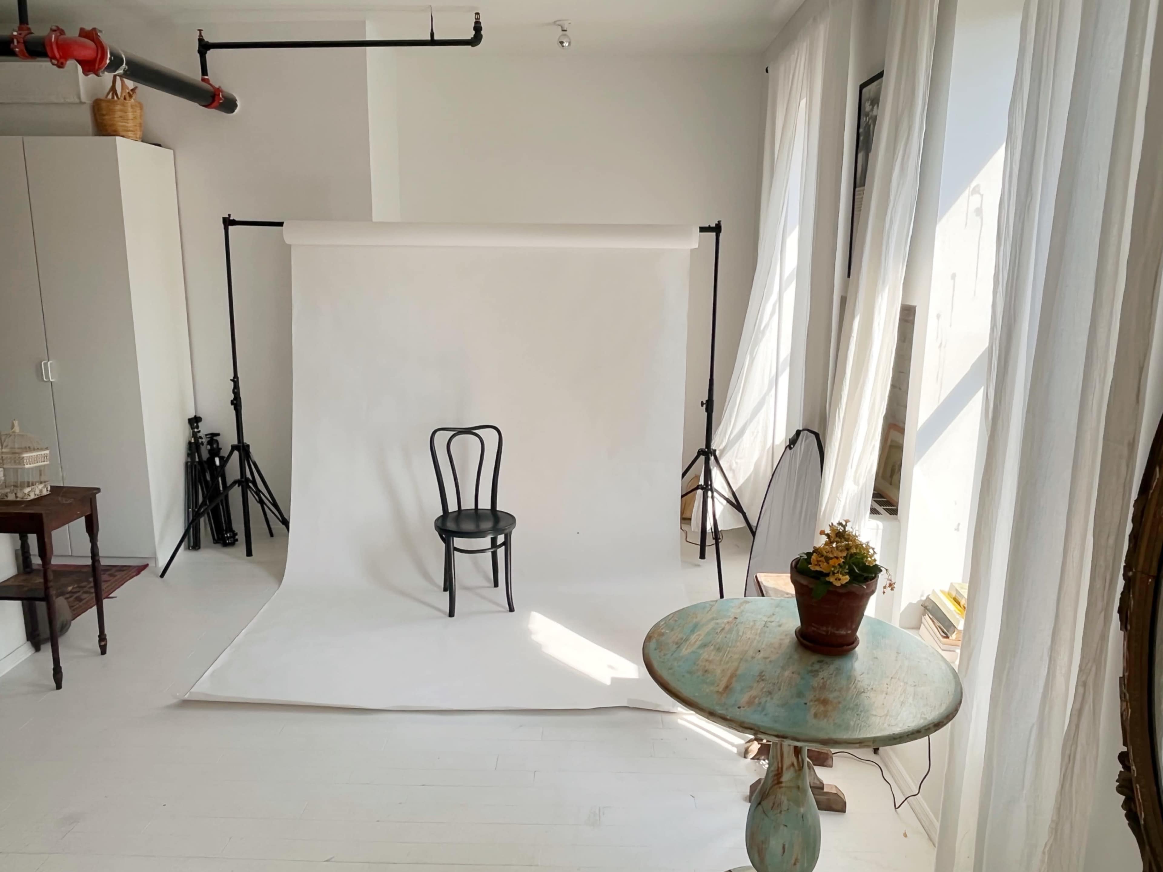 A photo studio setup with a black chair positioned on a white backdrop, surrounded by natural light and minimal furniture.