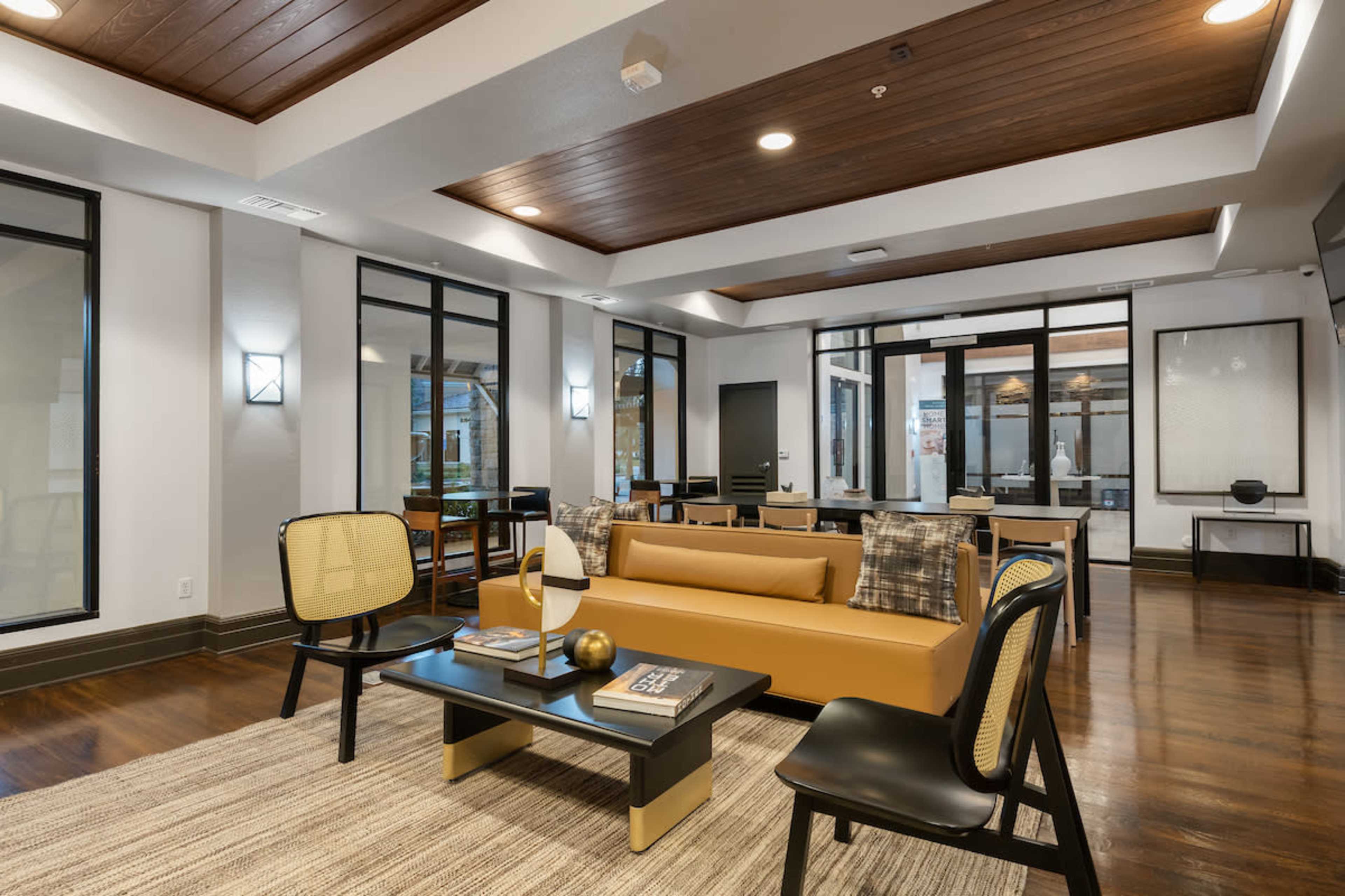 A modern lounge area with a brown sofa, black chairs, a glass coffee table, and large windows allowing natural light to enter.