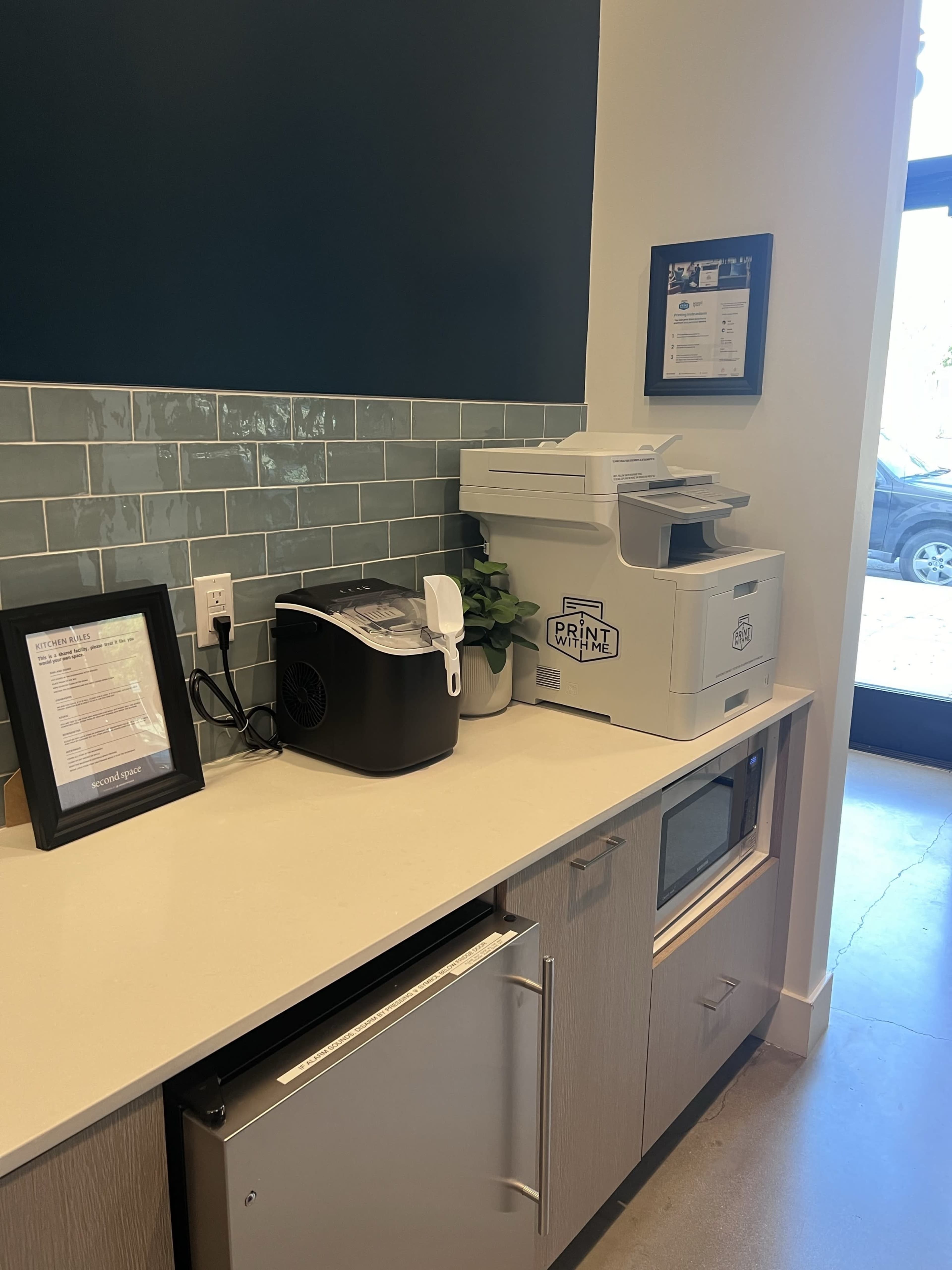 The image shows a kitchen area featuring a countertop with a coffee maker, a printer, and a small microwave, alongside a framed menu on the wall.