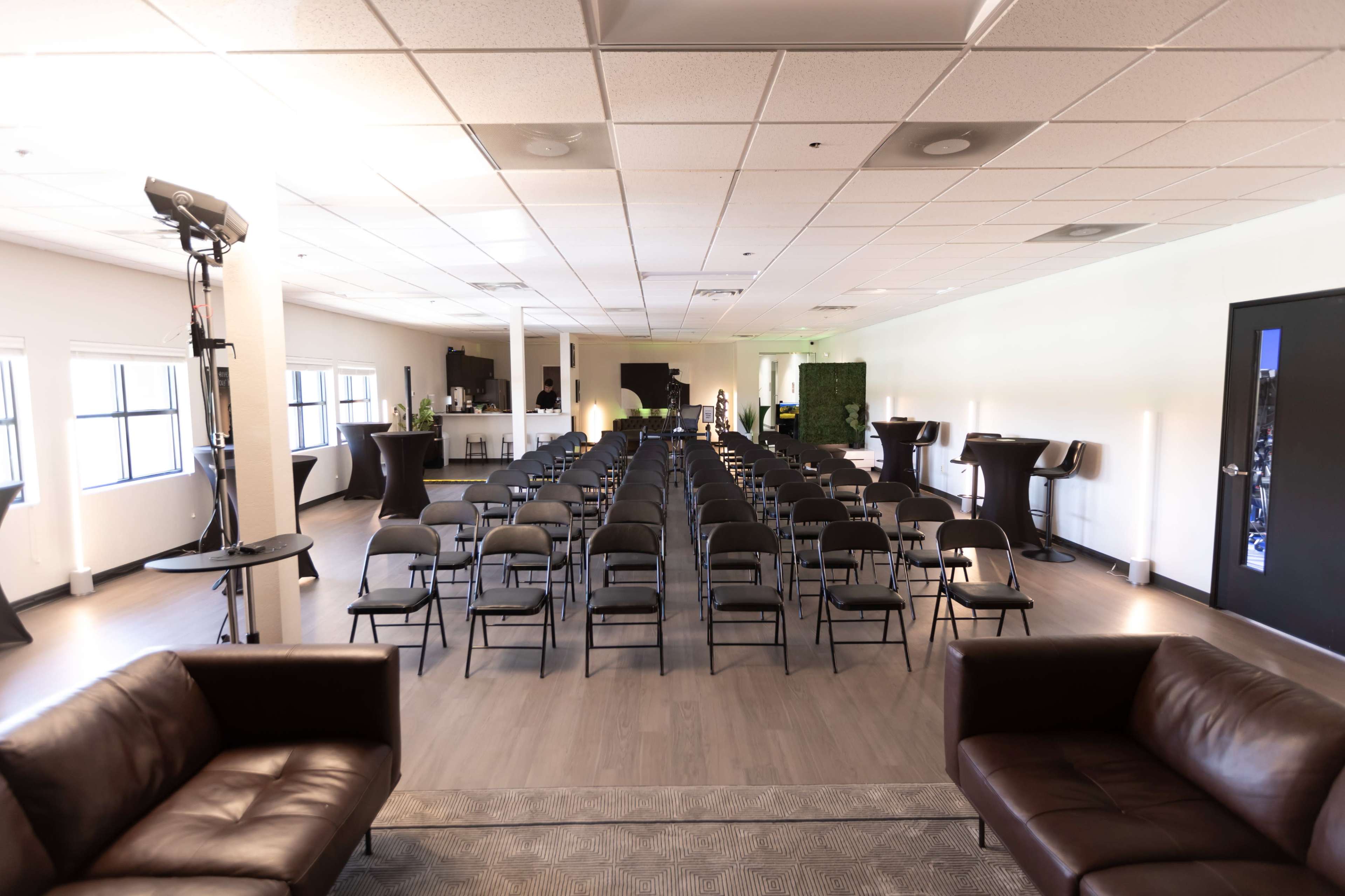 A spacious room arranged with rows of black chairs facing a presentation area, flanked by a bar and two leather couches.
