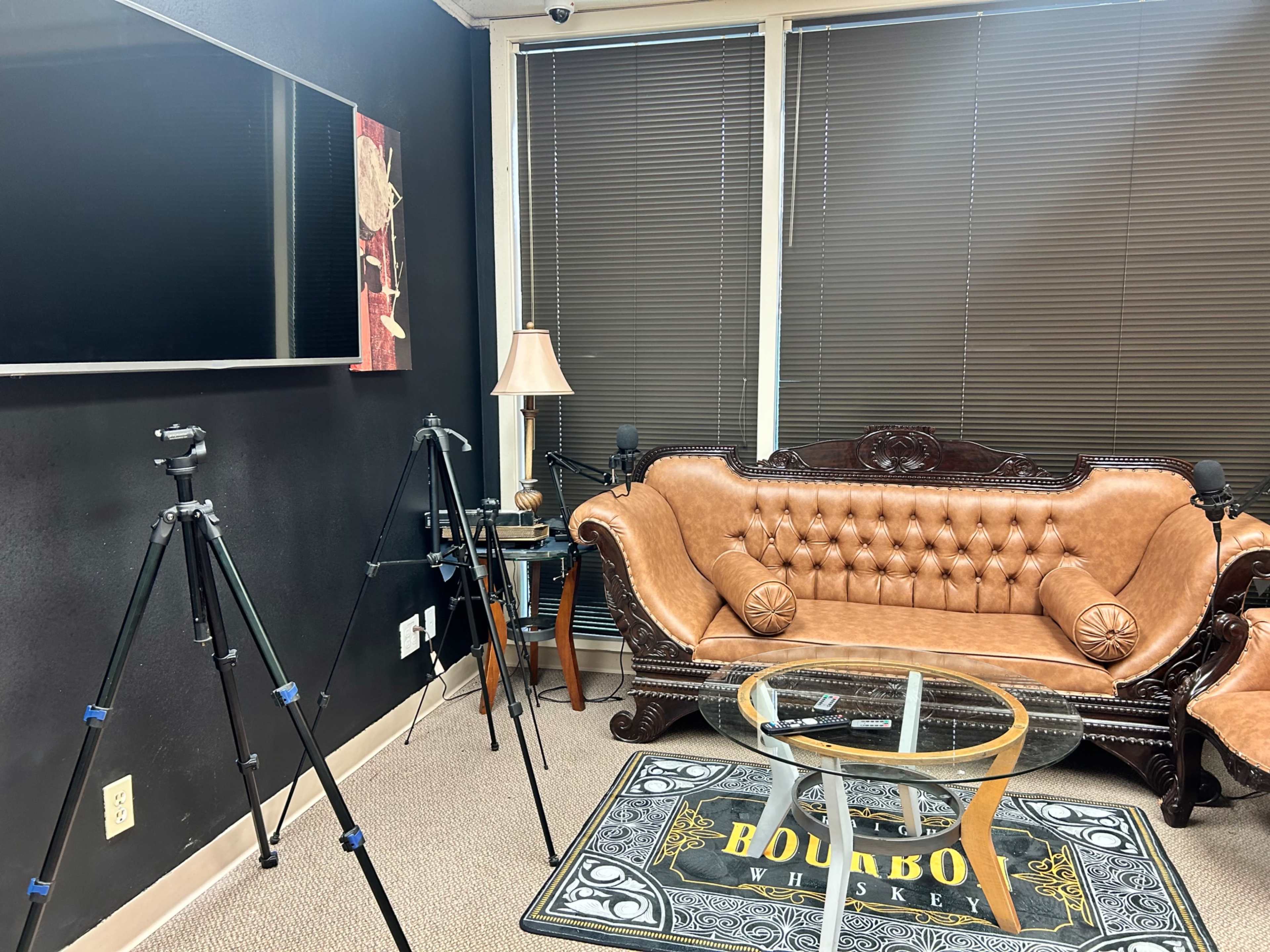 The scene features a well-furnished room with a brown leather sofa, a glass table, a carpet labeled "BOURBON WHISKEY," and several camera tripods positioned around the space.