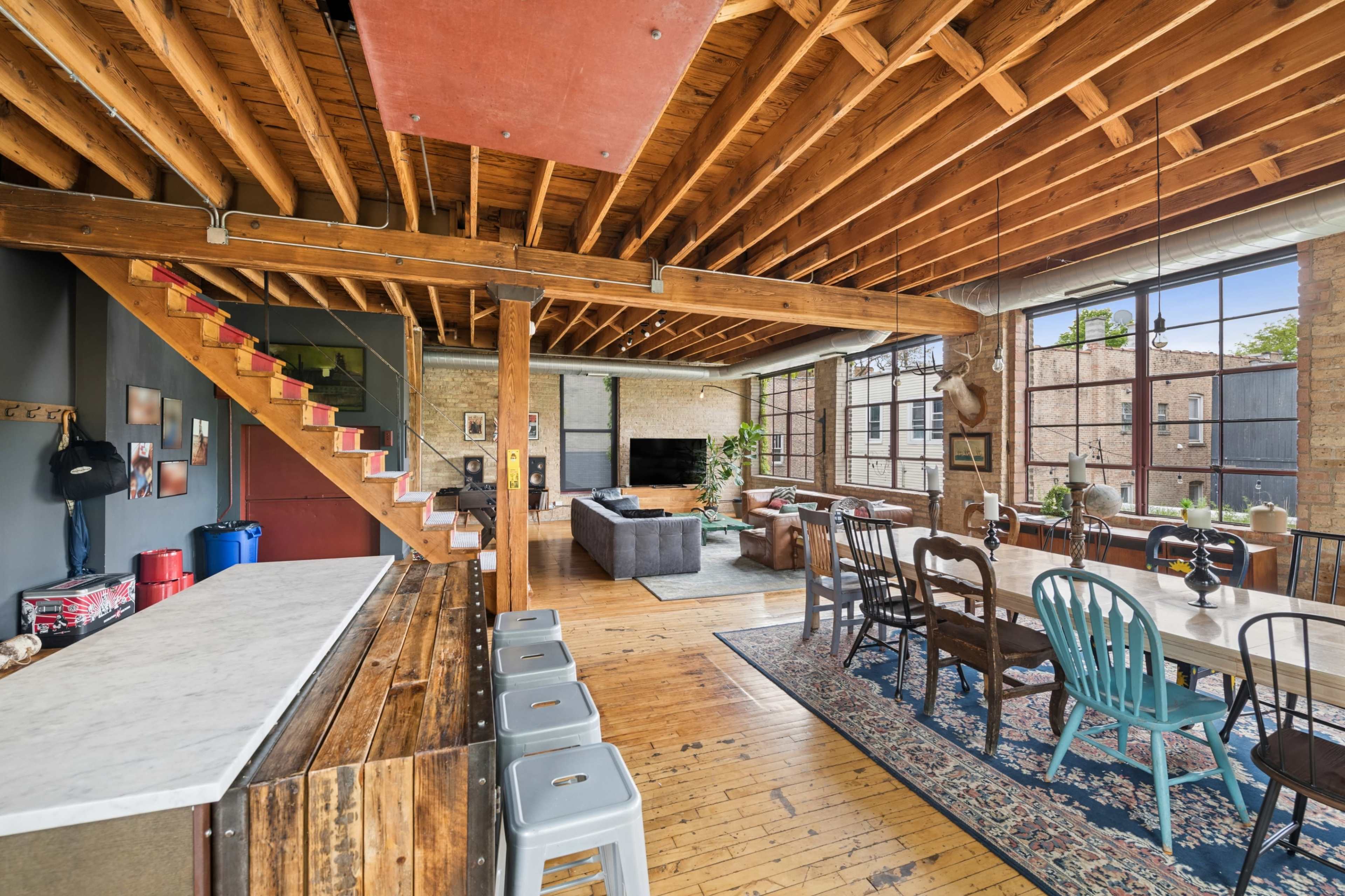The image shows a spacious, modern loft with wooden beams, a large seating area, and a dining table situated near wide windows.