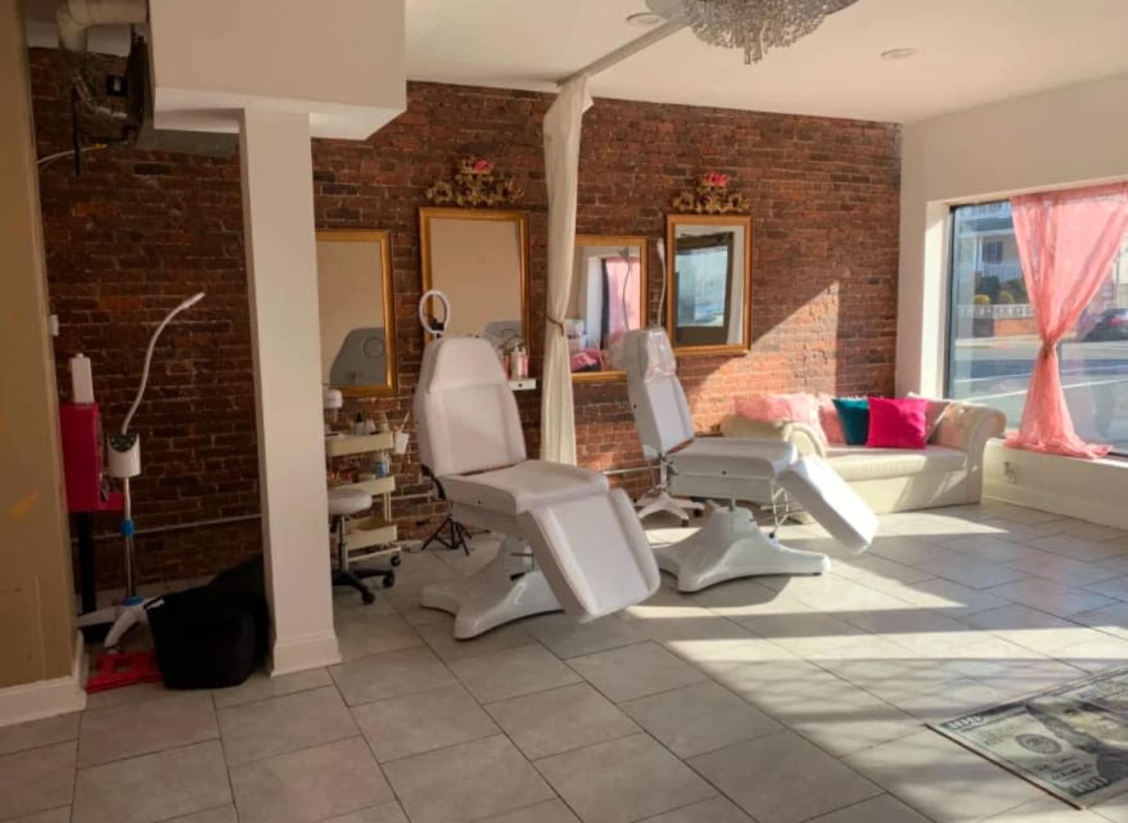 A well-lit beauty treatment room featuring two white treatment chairs, a sofa, and large windows with sheer curtains, against a backdrop of exposed brick walls and decorative mirrors.