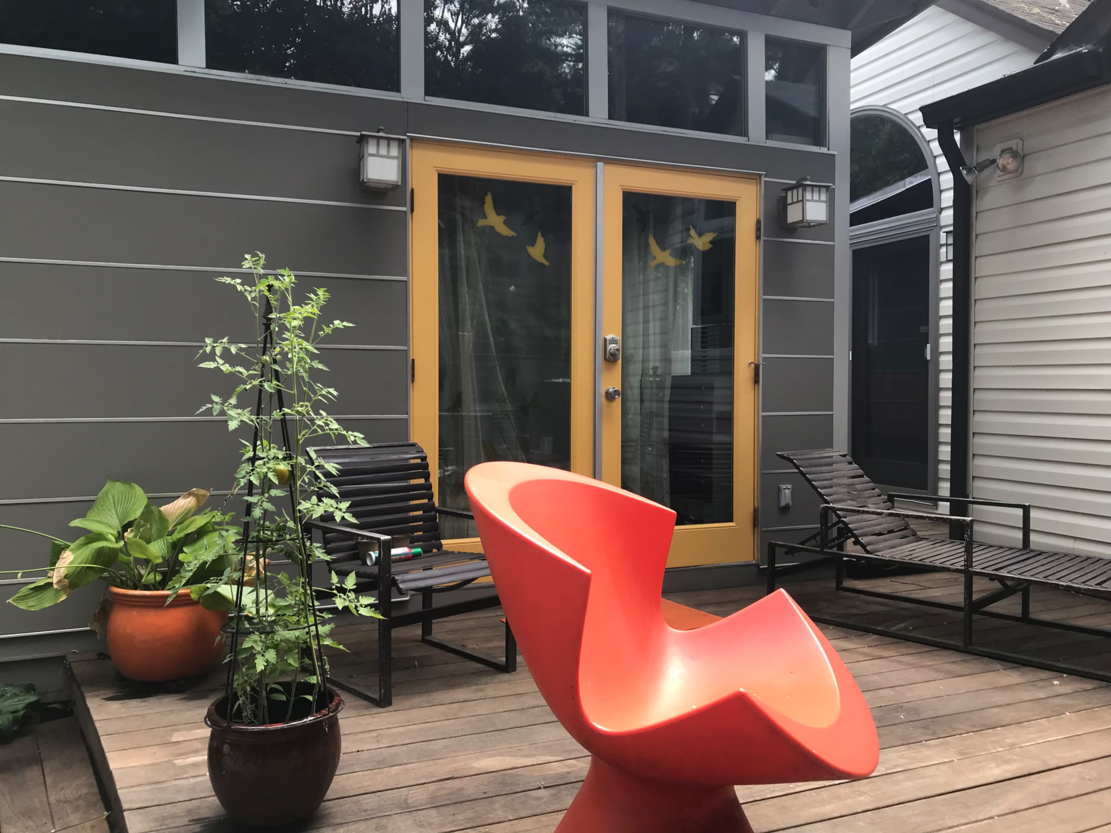 A deck area with a bright orange chair, two lounge chairs, potted plants, and a set of yellow doors leading into the house.