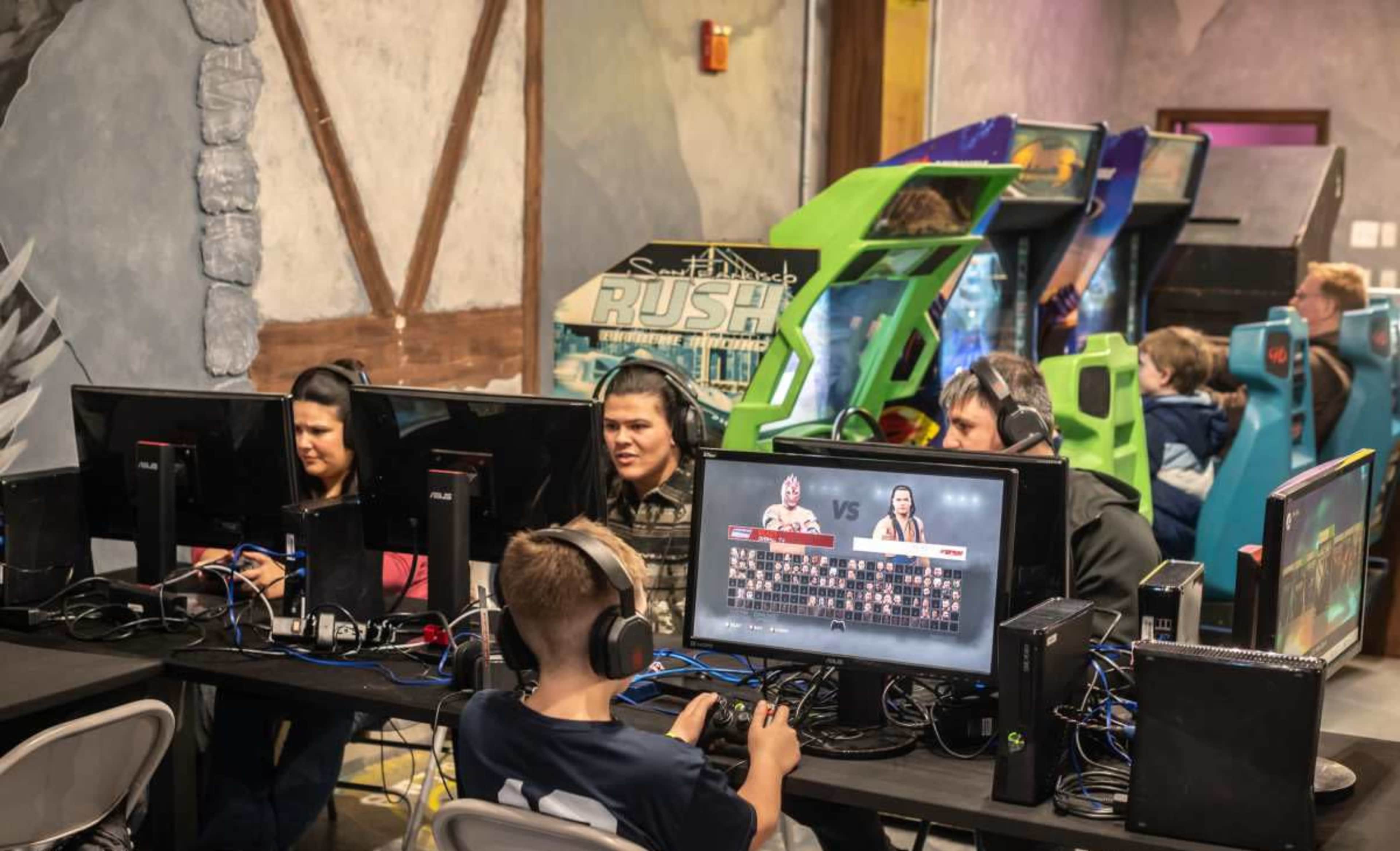 A group of people, including children and adults, is seated at computers playing video games in a lively gaming center filled with arcade machines in the background.
