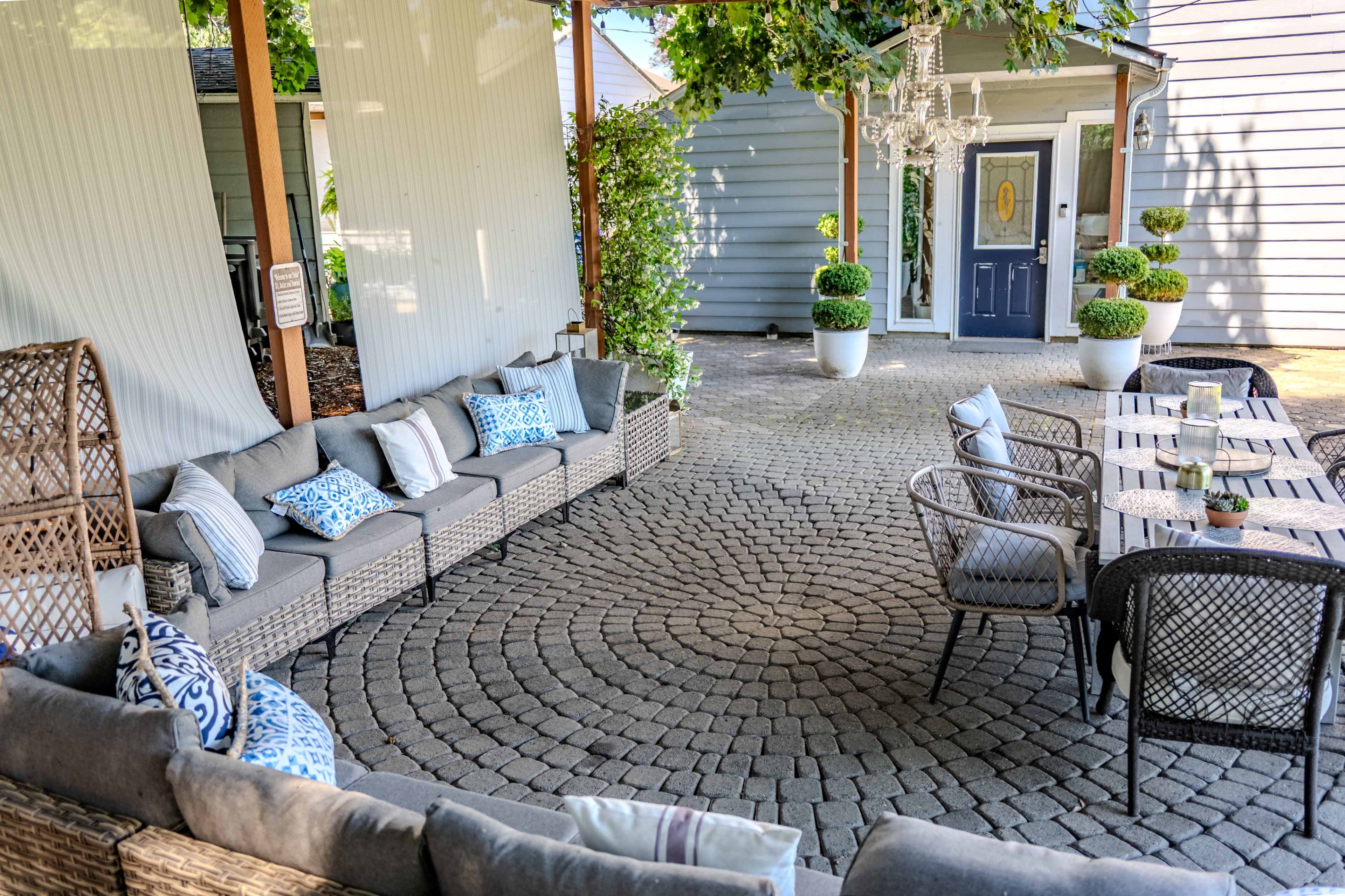 The image shows a cozy outdoor seating area arranged with a circular stone pattern, featuring a long sofa, tables, and landscaped greenery.
