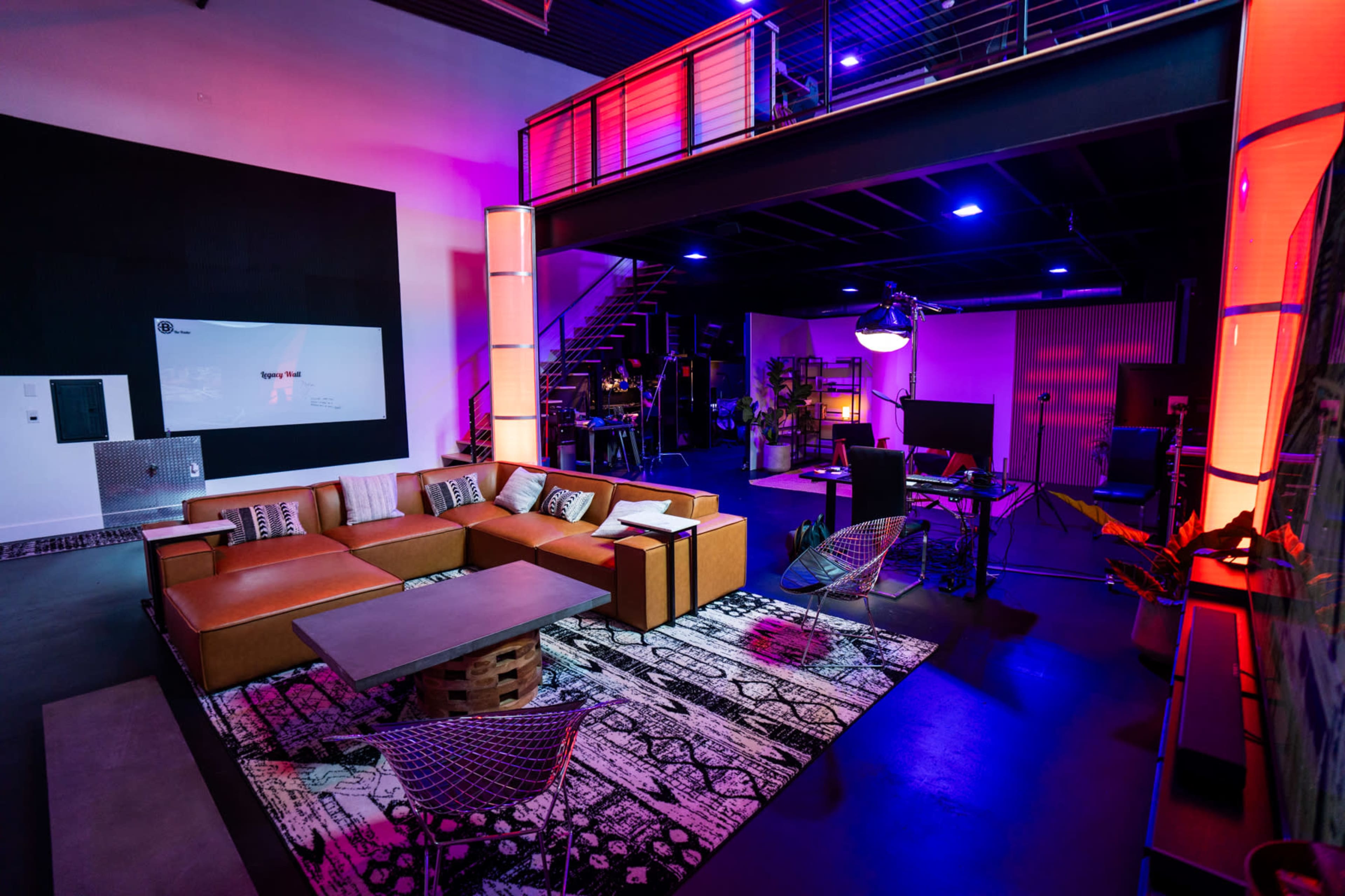 The image depicts a modern, open workspace featuring a lounge area with leather sofas, a coffee table, and decorative rugs, illuminated by colorful lighting.