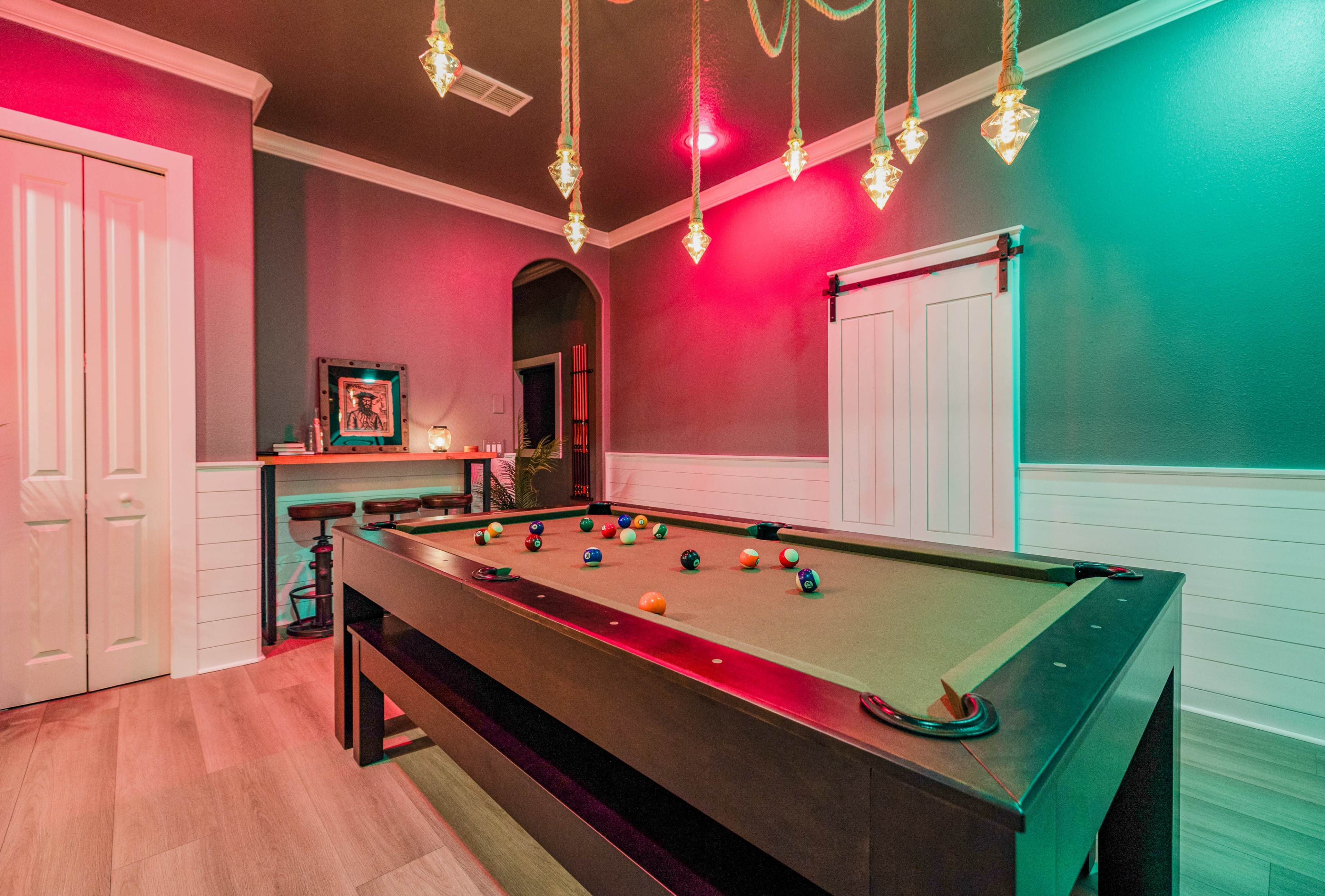 The image shows a billiards room with a pool table, illuminated by decorative pendant lights and colored wall lights, featuring a bar area and a door in the background.