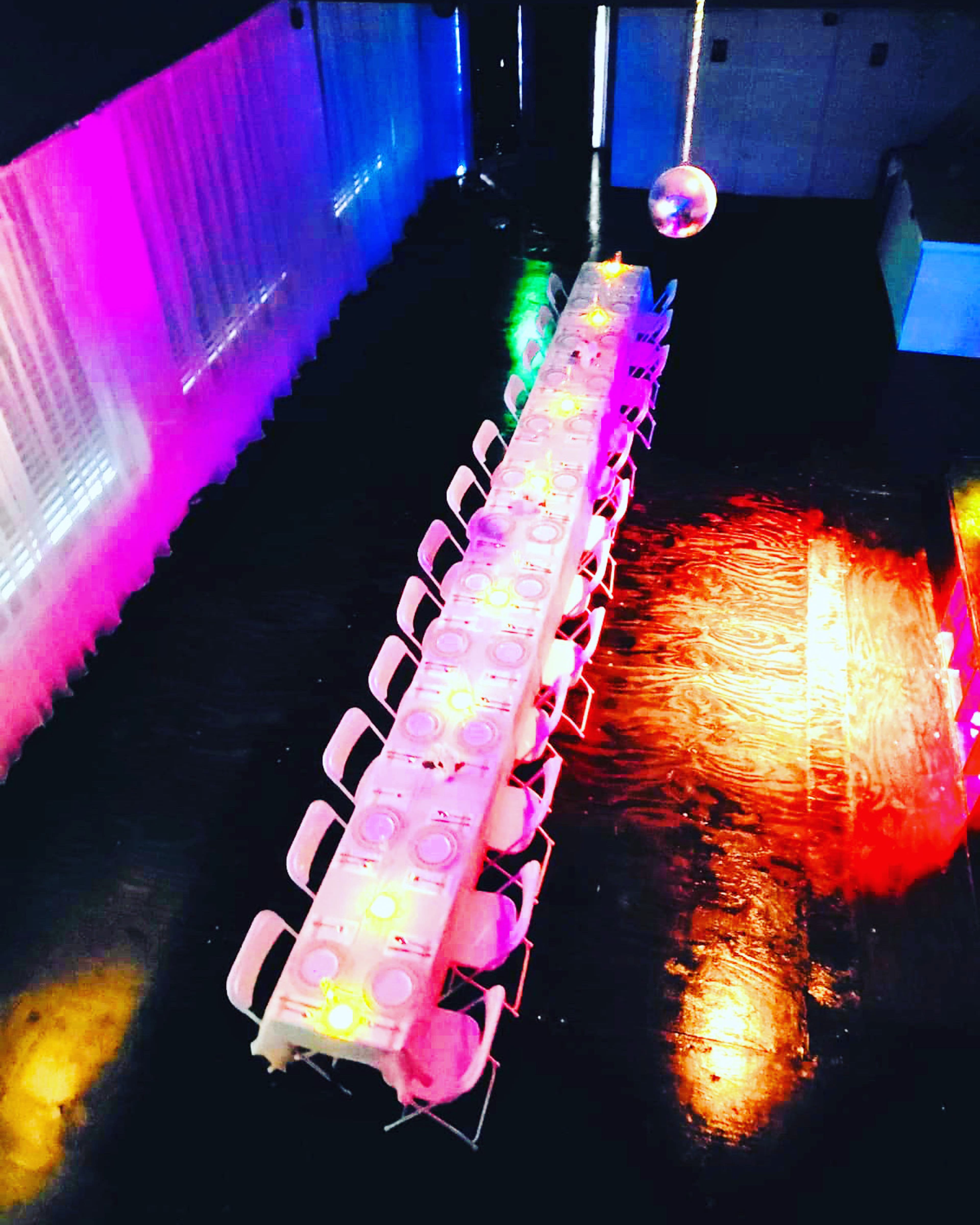 A long, rectangular dining table is set with plates and illuminated by colorful lights in a dimly lit venue.
