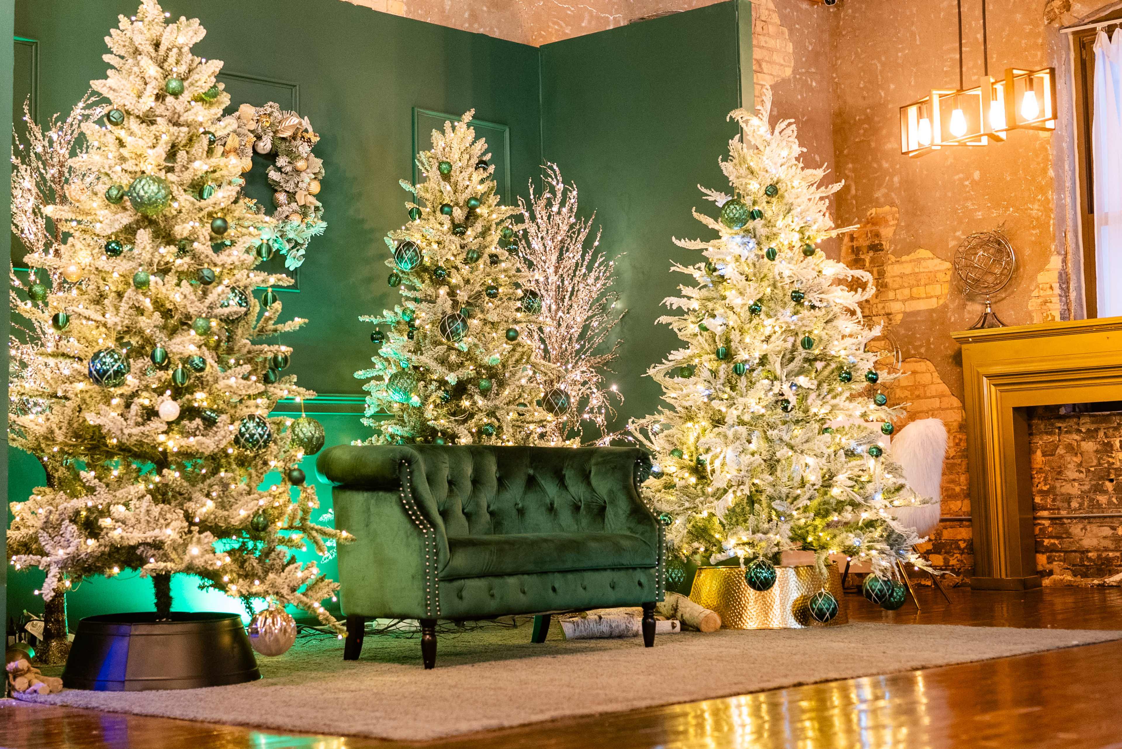 The scene features three decorated Christmas trees surrounded by a cozy green velvet sofa in a festive interior setting.