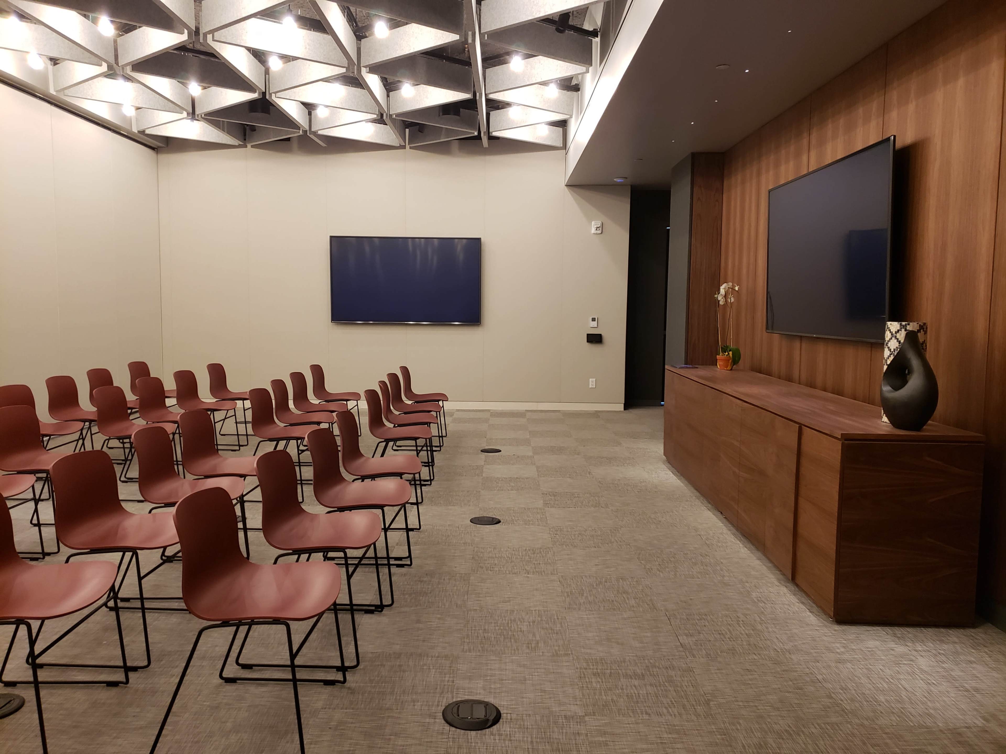 A conference room features rows of red chairs facing a wooden media console with two wall-mounted screens.