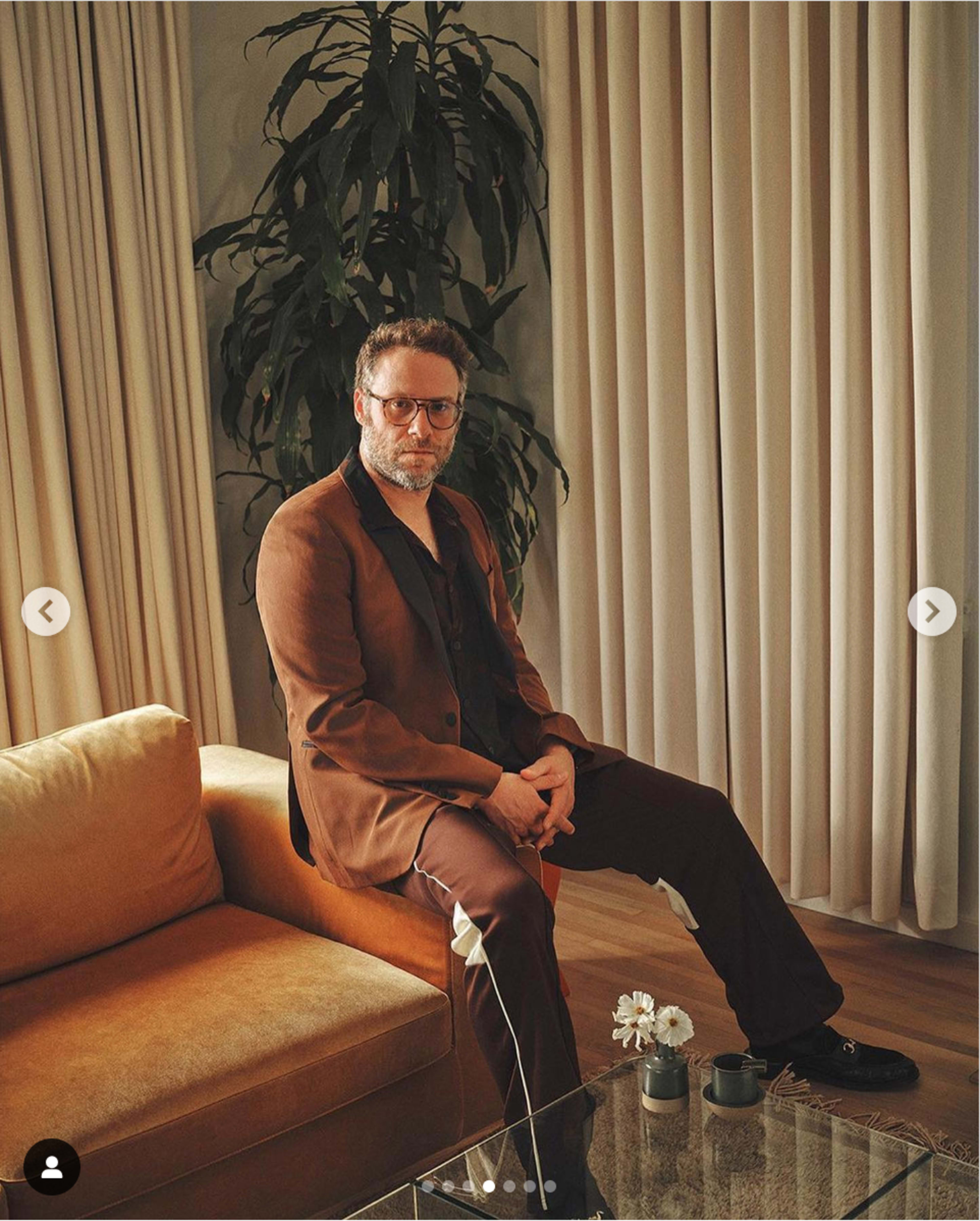A man wearing a brown suit sits on a couch in a room with a large potted plant and curtains.