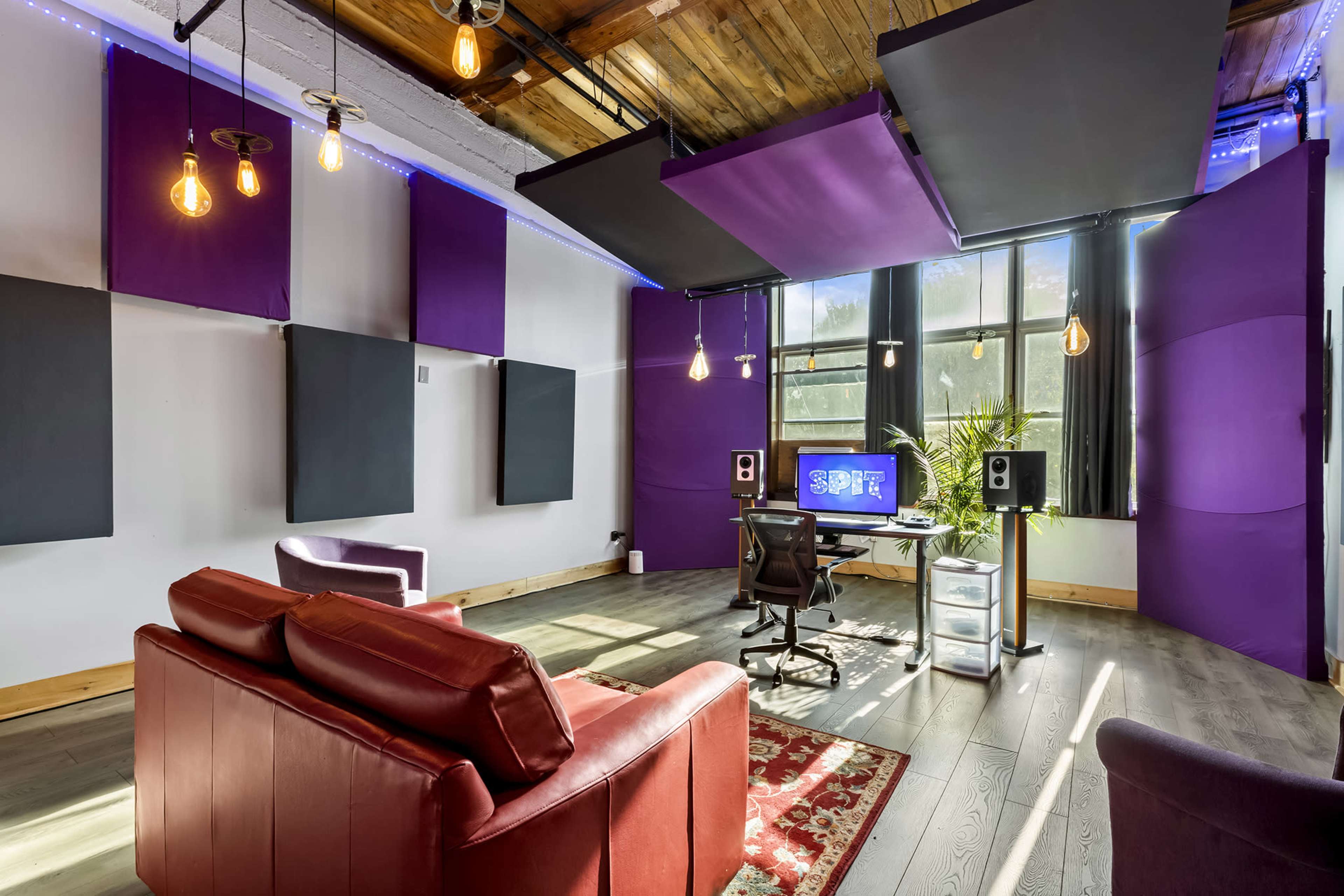 A modern studio space with purple acoustic wall panels, a desk with a computer setup, and a seating area featuring a red couch and a decorative rug.