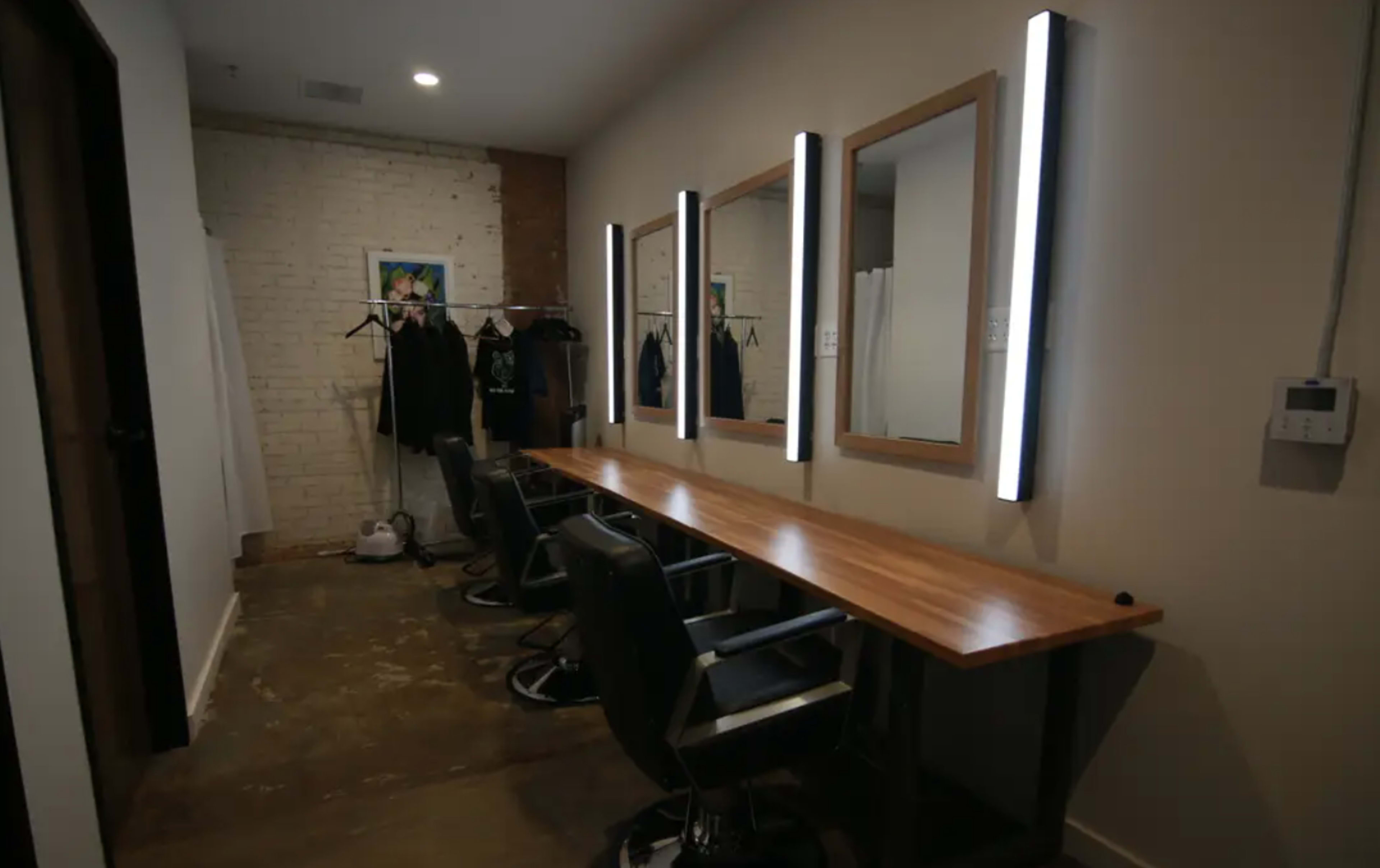 The image shows a salon interior with three illuminated mirrors above wooden stations and black salon chairs, alongside a clothes rack on the wall.