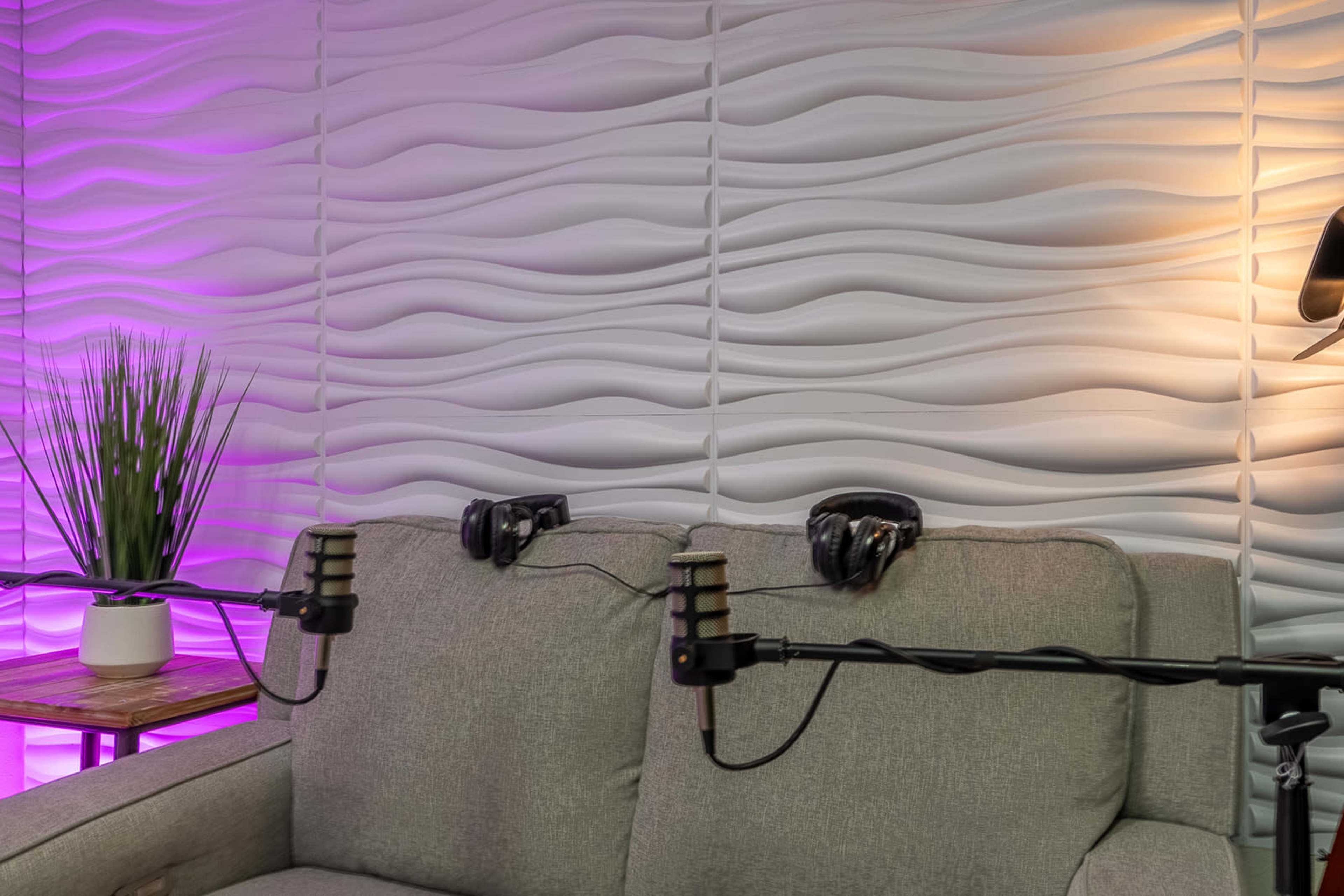 A light gray sofa with headphones resting on its back is positioned near a textured wall illuminated in purple light.