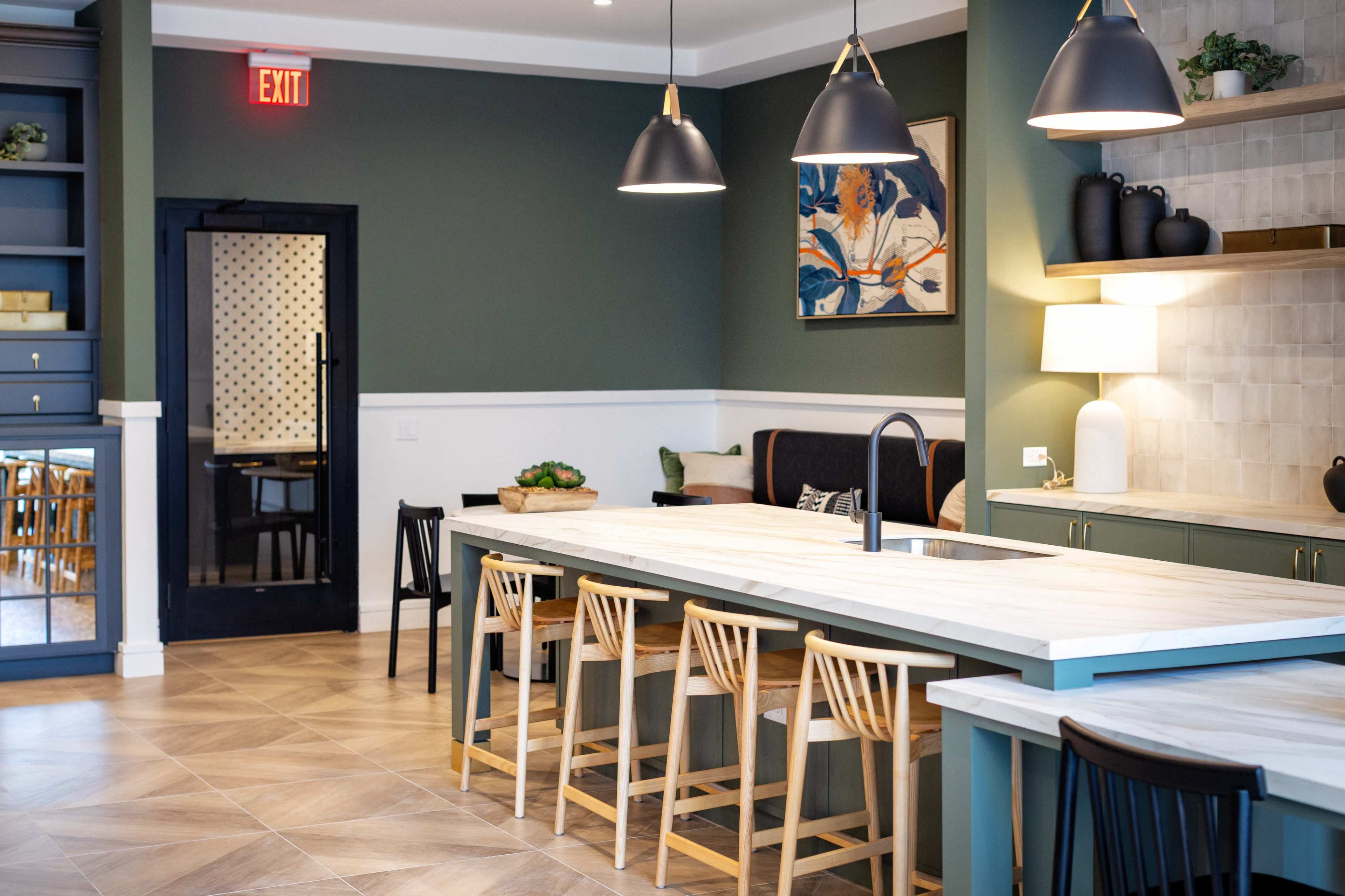 The image shows a modern kitchen space with a large island, wooden bar stools, and contemporary light fixtures, featuring green and neutral tones on the walls and cabinetry.