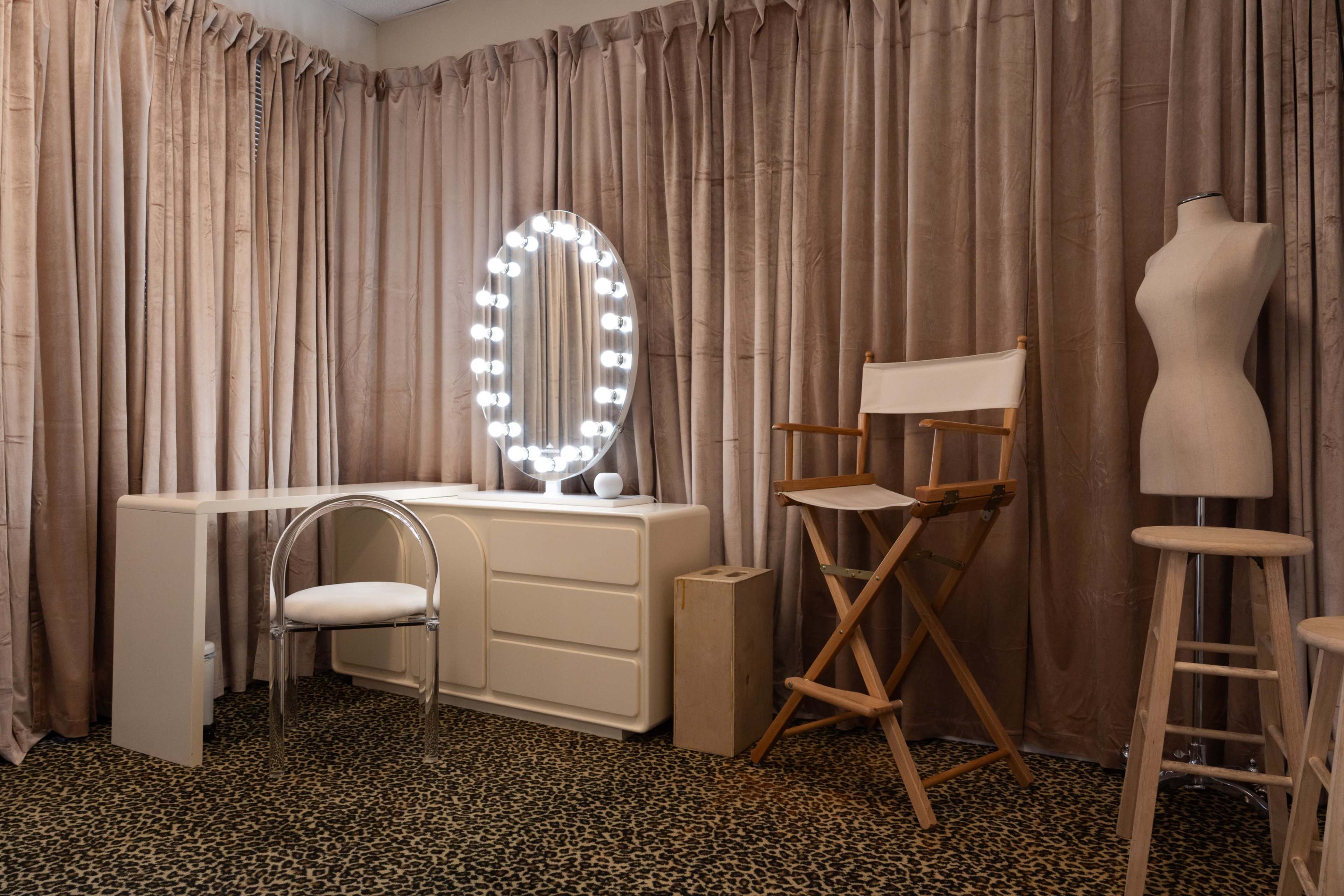 The image shows a dressing room with a makeup vanity illuminated by a circular mirror, a wooden director's chair, and a dress form against a backdrop of beige curtains and a leopard print carpet.
