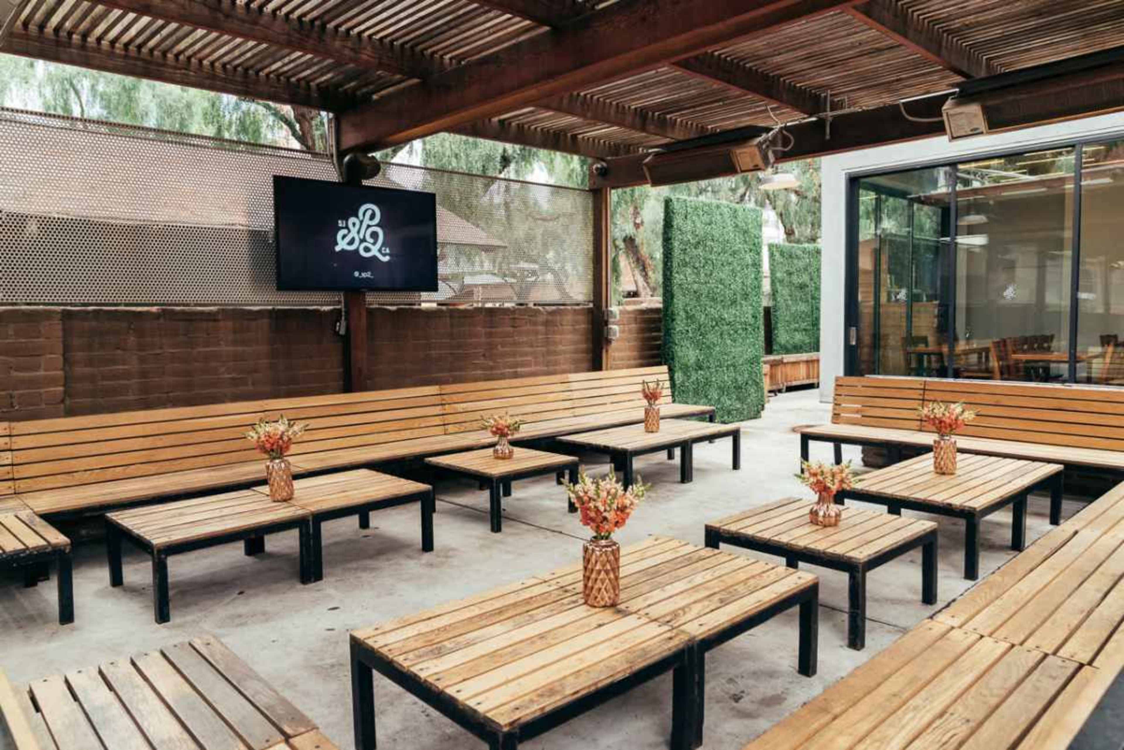 The image shows a spacious outdoor seating area with wooden benches and tables arranged neatly, adorned with vases of flowers on each table, and a screen mounted on the wall.