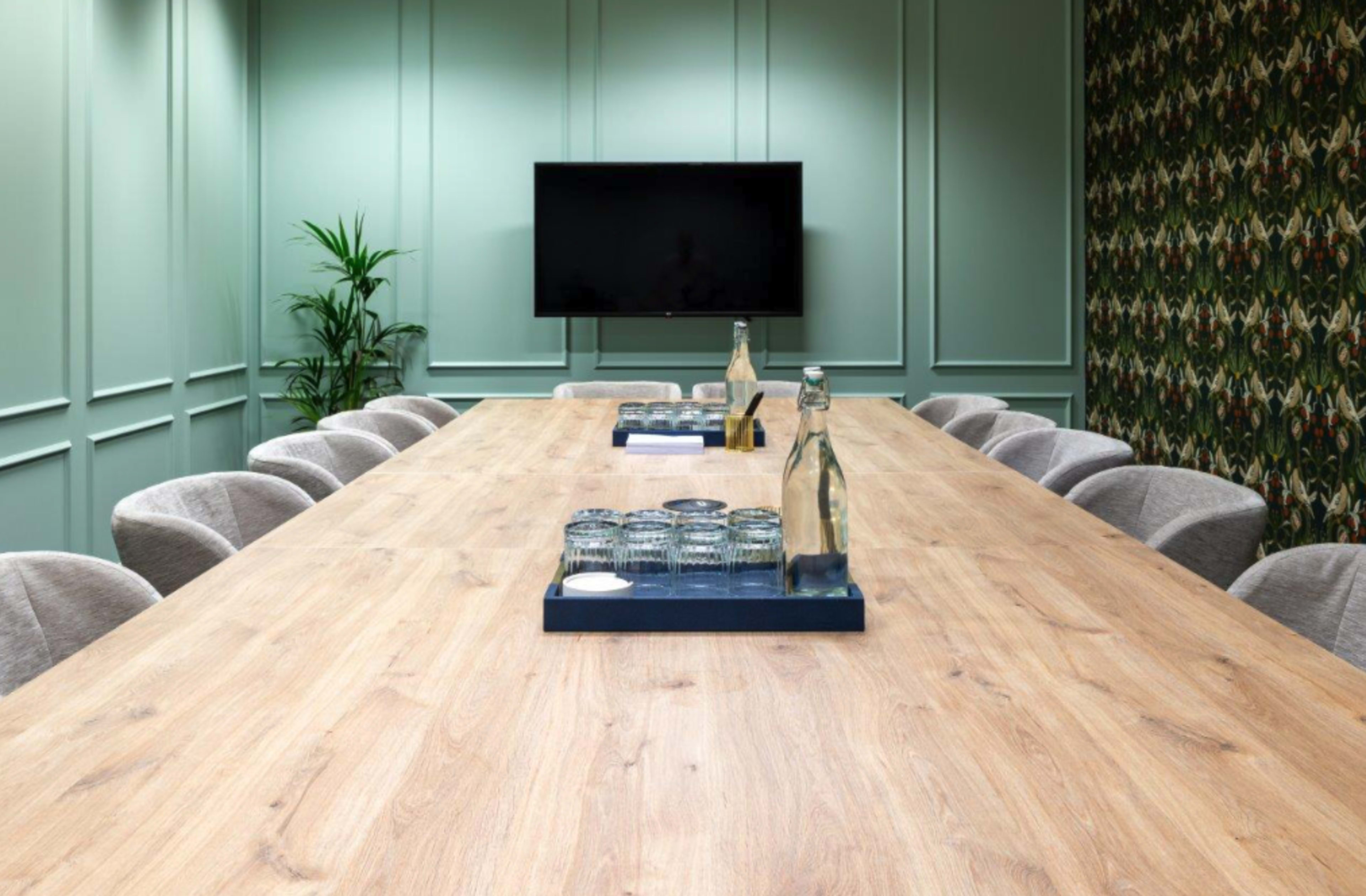 A large wooden conference table is set with glasses and water bottles in a green-paneled meeting room with a television screen and patterned wallpaper.