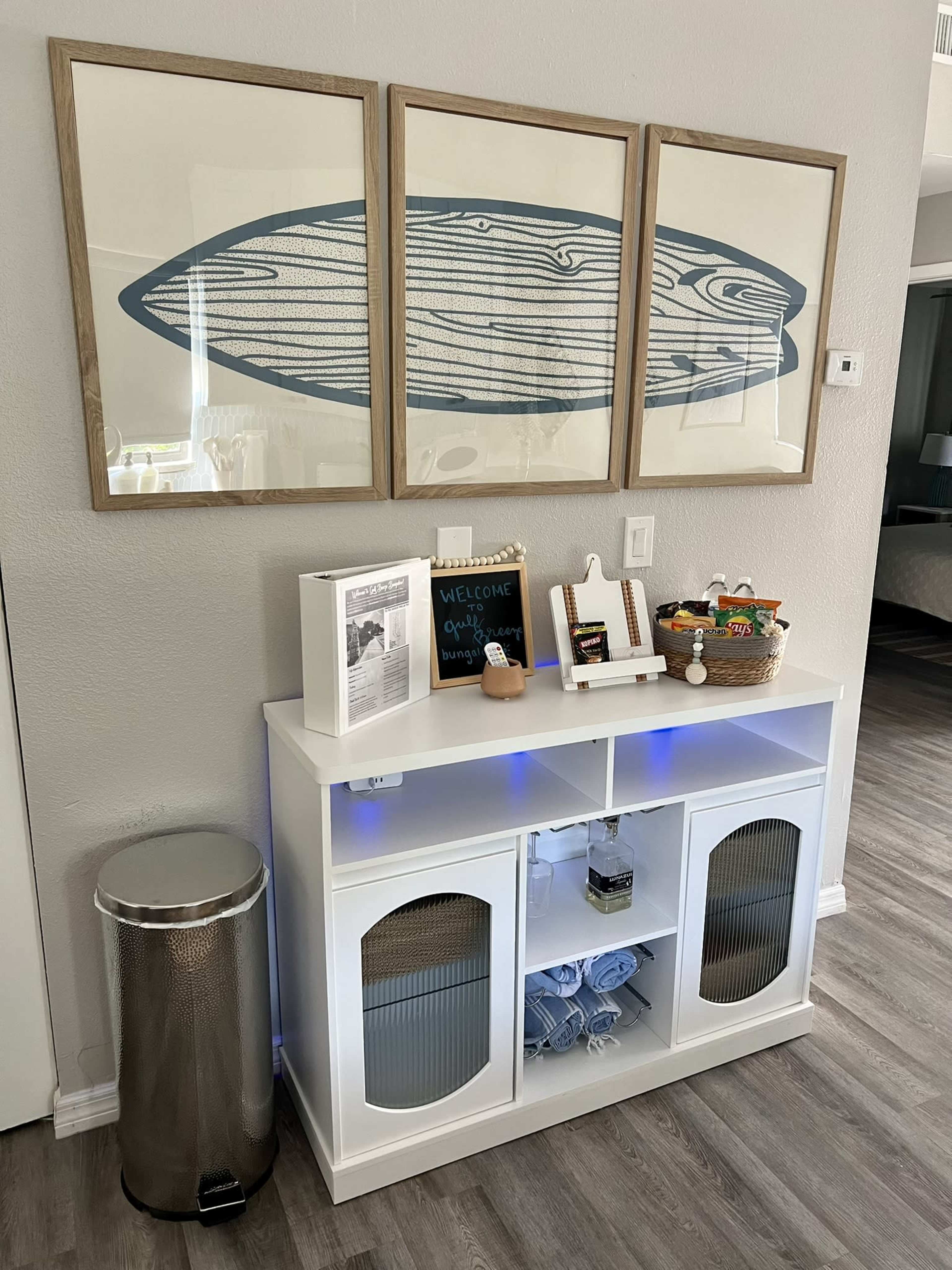 The image shows a modern entryway featuring a white console table with a blue-lit shelf, adorned with decorative items, a welcome sign, and three framed artworks above it.