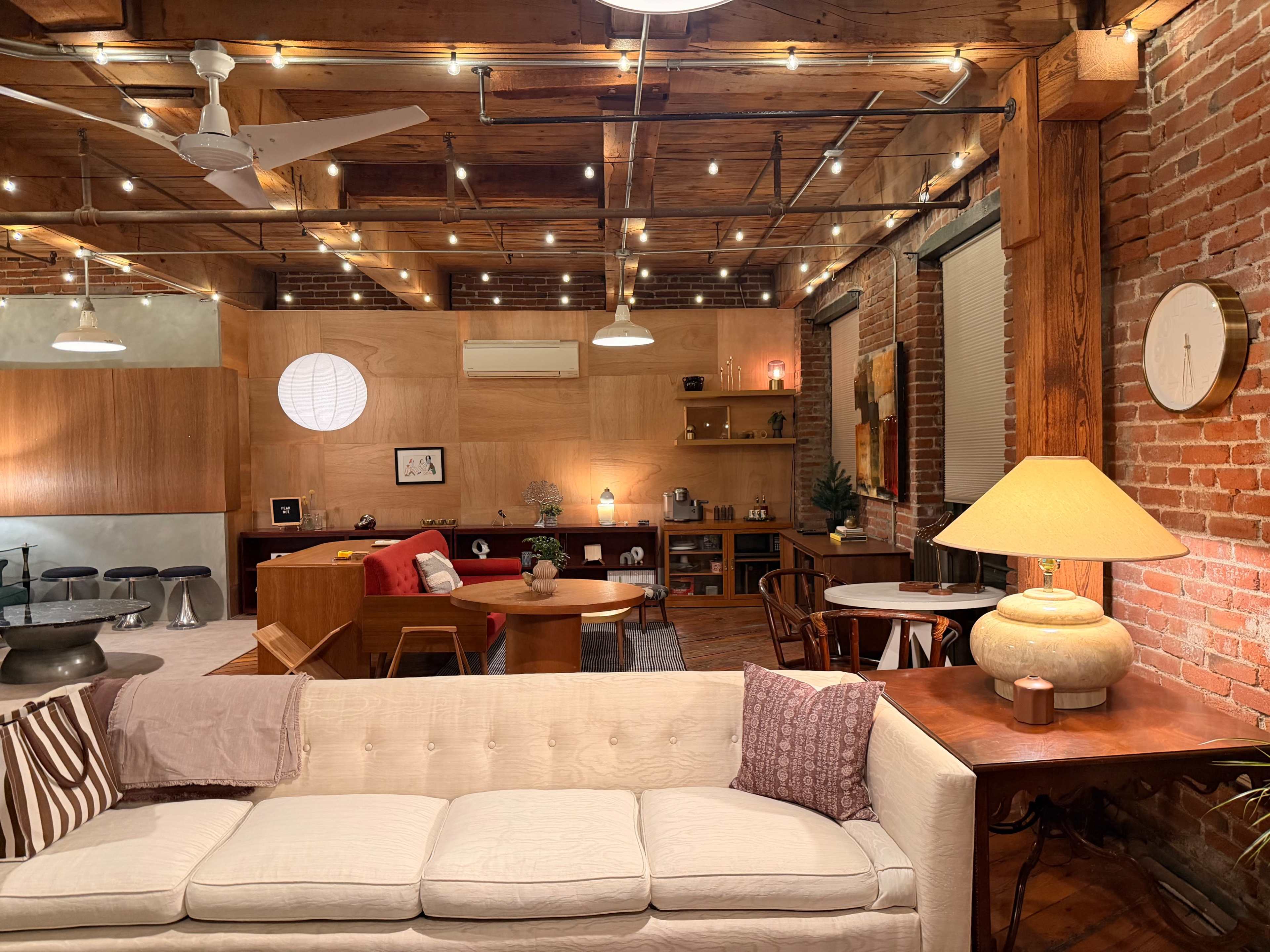 The image shows a spacious, well-lit living area with exposed brick walls, wooden beams, and various furniture arrangements, including a white sofa, a wooden dining table, and decorative lighting.