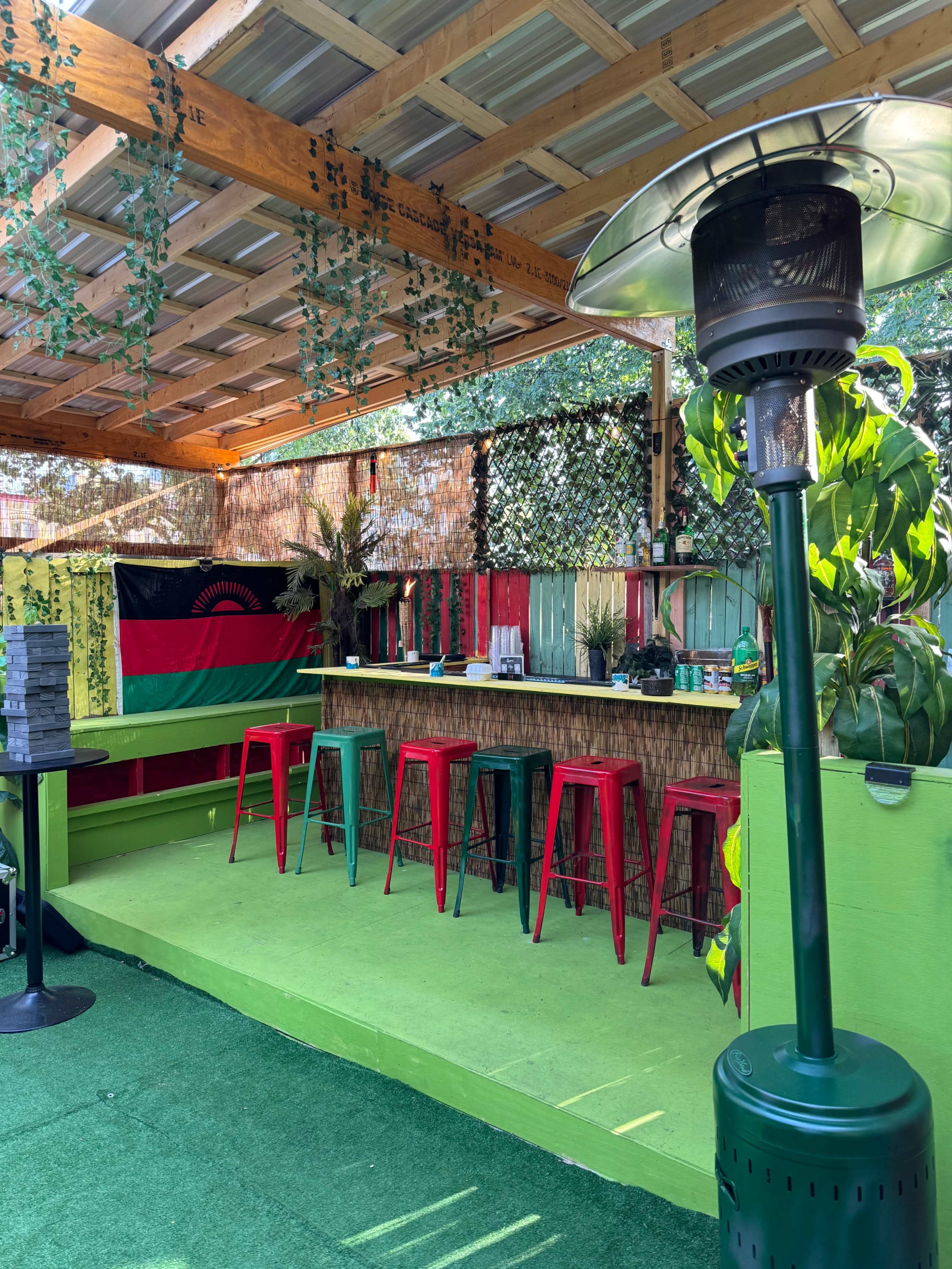 The image shows a vibrant outdoor bar area with green flooring, red and green tall stools, and a thatched roof, decorated with plants and a flag in the background.