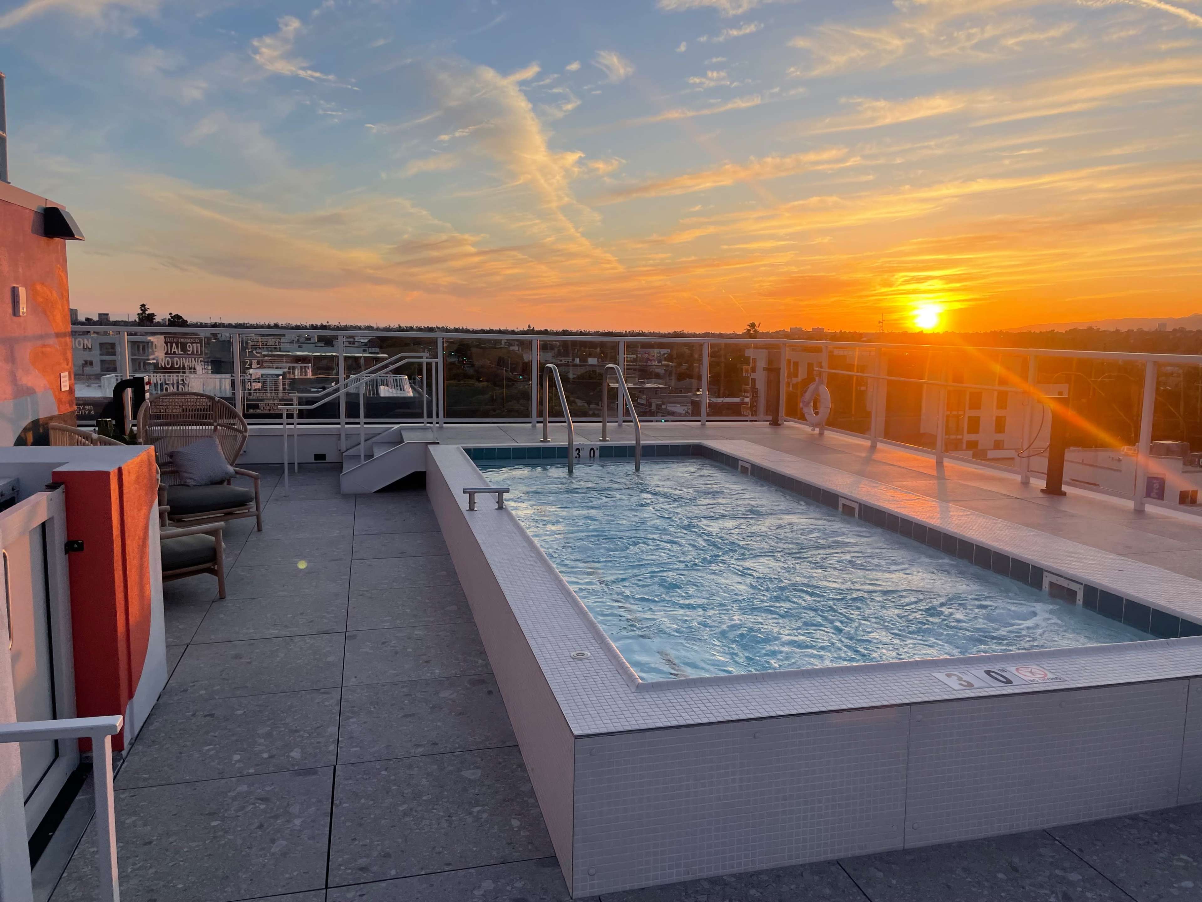 A rooftop pool with clear water reflects the vibrant colors of a sunset over a city skyline.