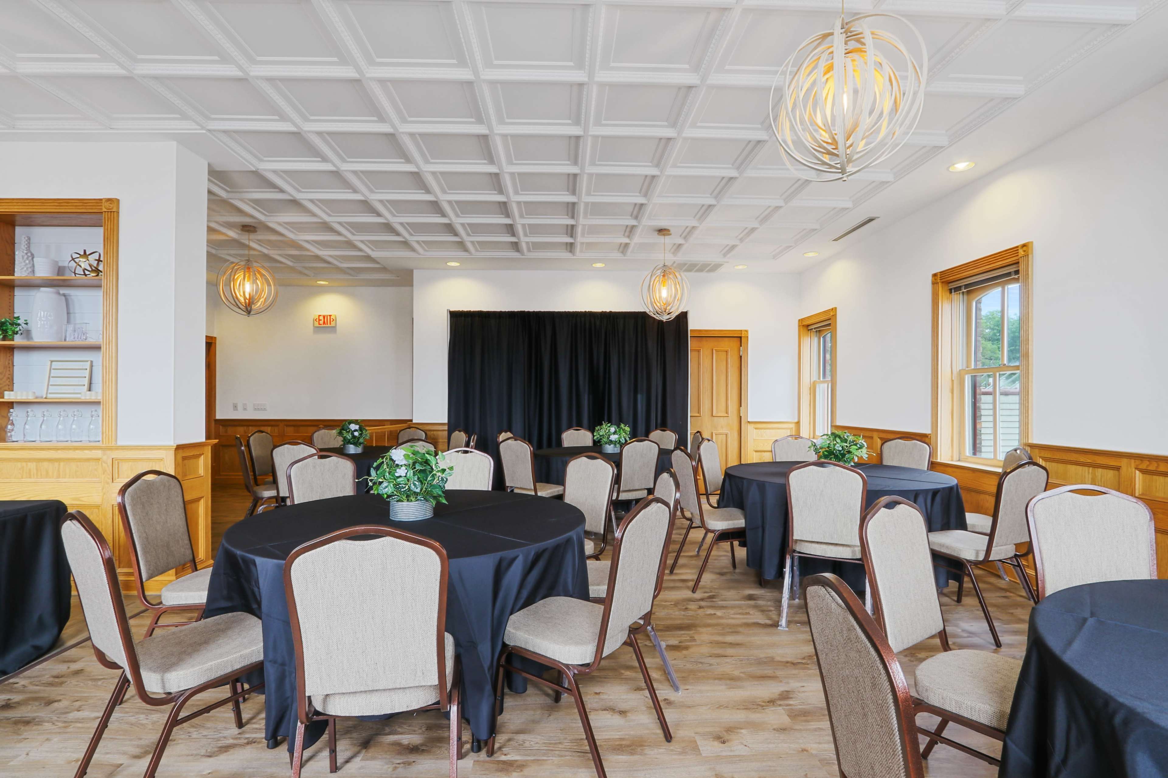 A banquet room features several round tables with black tablecloths and beige chairs, arranged around a stage draped in black fabric.