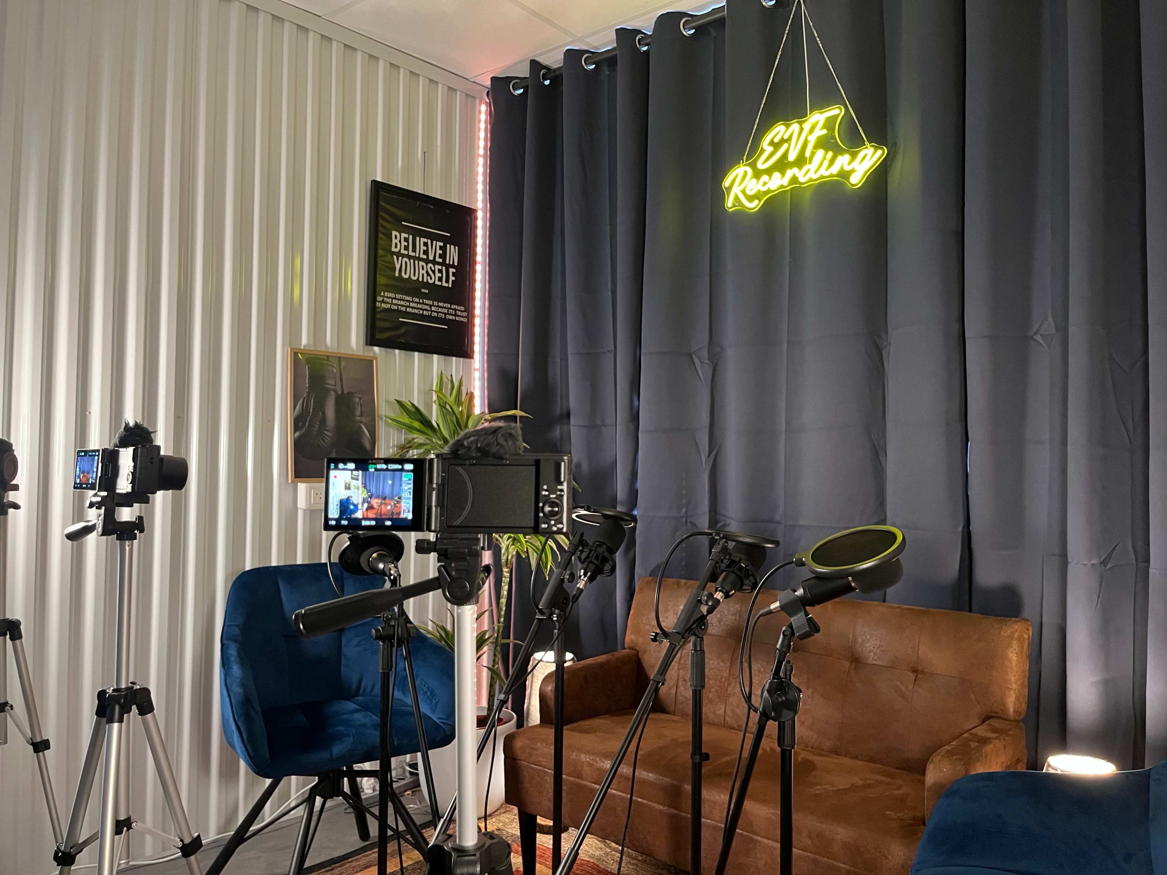 The image shows a recording studio setup featuring a brown leather couch, multiple cameras on tripods, microphones, and a neon sign that reads "EVT Recording" against a dark curtain backdrop.