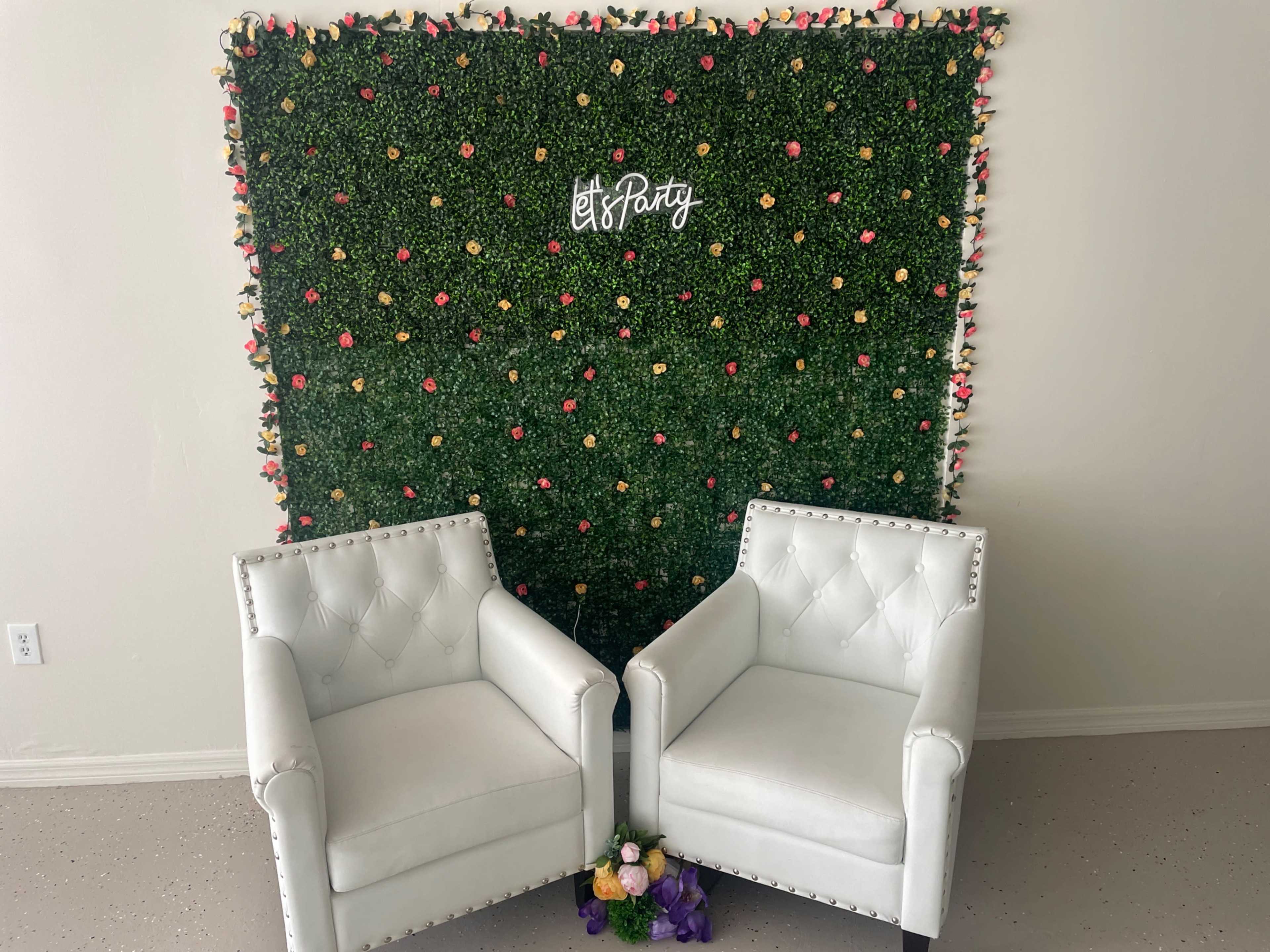 A backdrop of artificial greenery adorned with colorful flowers and the phrase "Let's Party" stands behind two white upholstered chairs in a bright room.