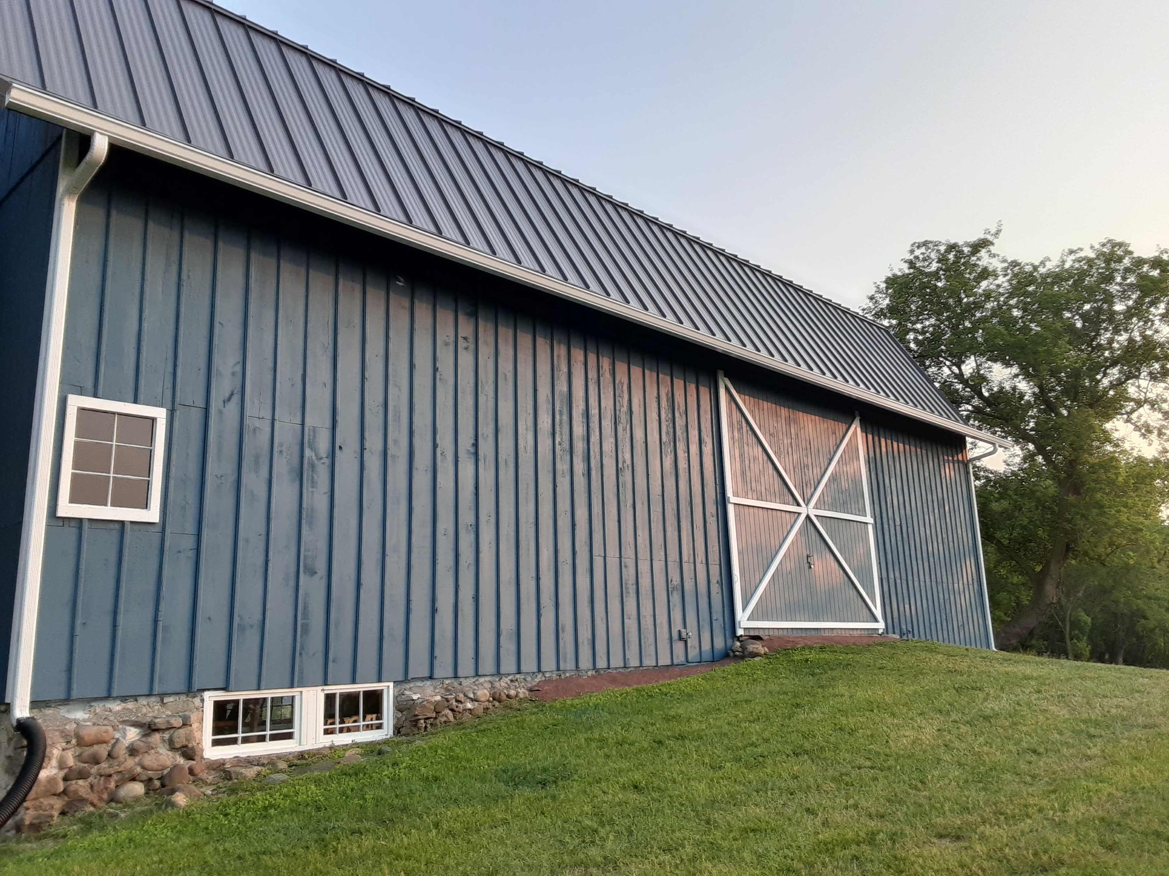The image shows a blue barn with a metal roof, set against a grassy hillside and trees in the background.