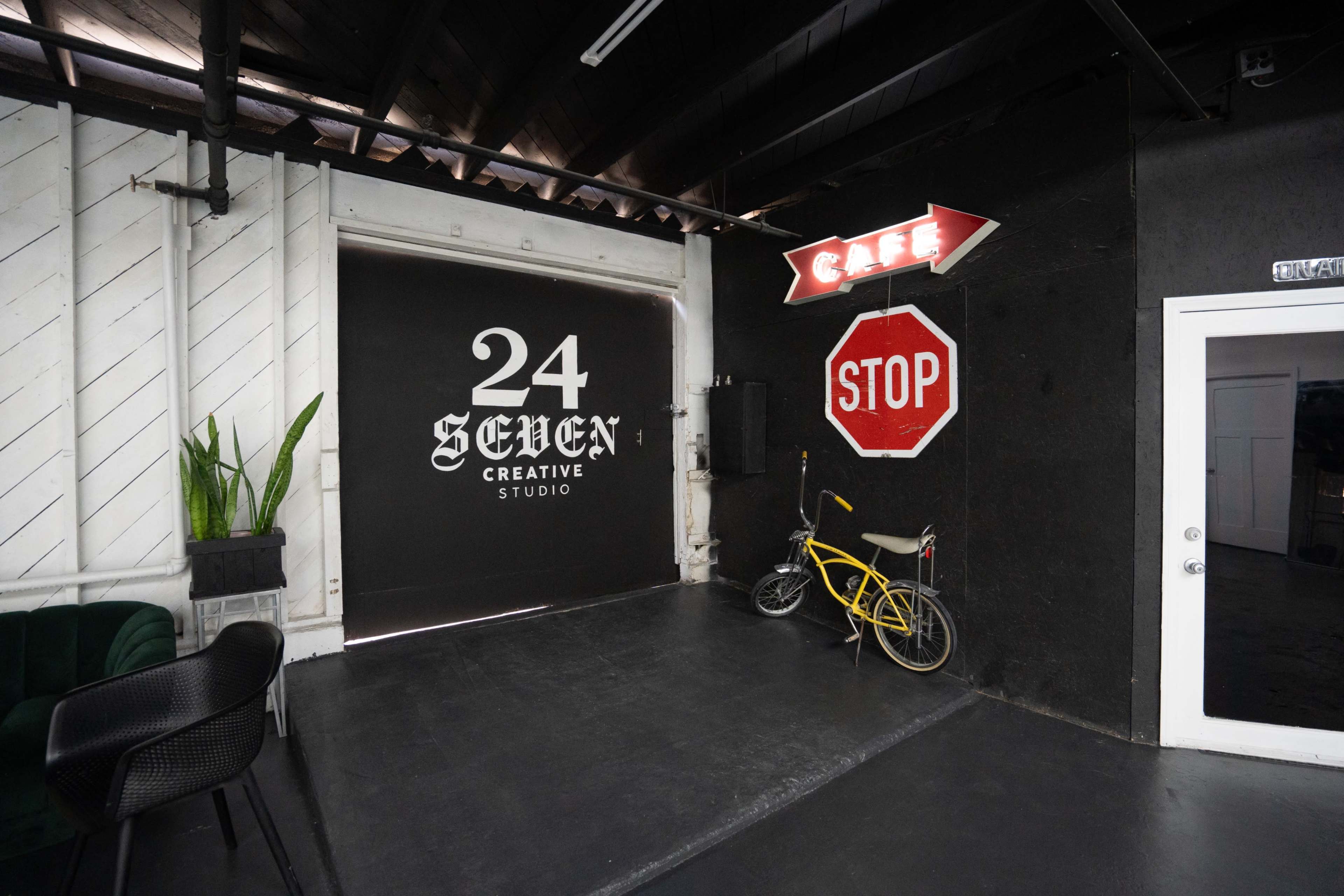 The image shows a creative studio entrance featuring a large black garage door with the text "24 SEVEN CREATIVE STUDIO," along with a café sign and a bicycle leaning against the wall.