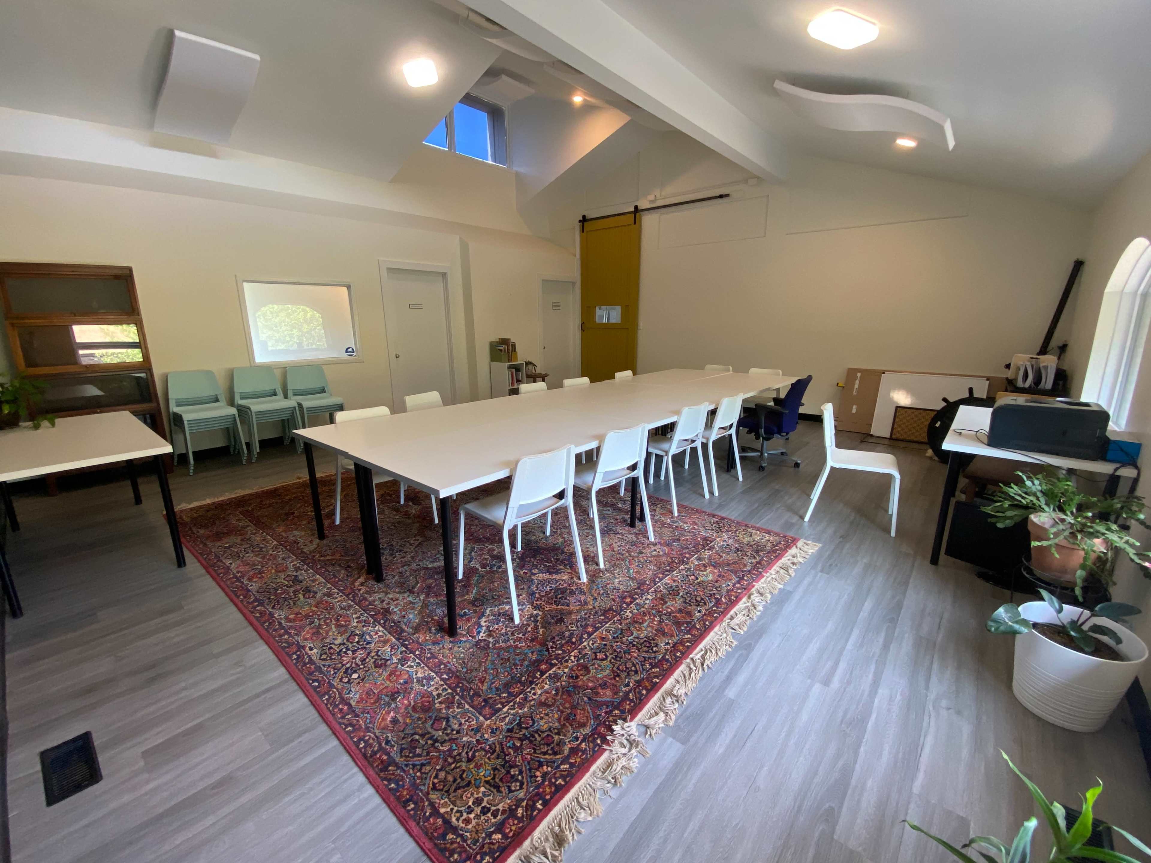 The image shows an interior room set up with several tables and chairs arranged around a large area rug, with a shelf and a few plants in the background.