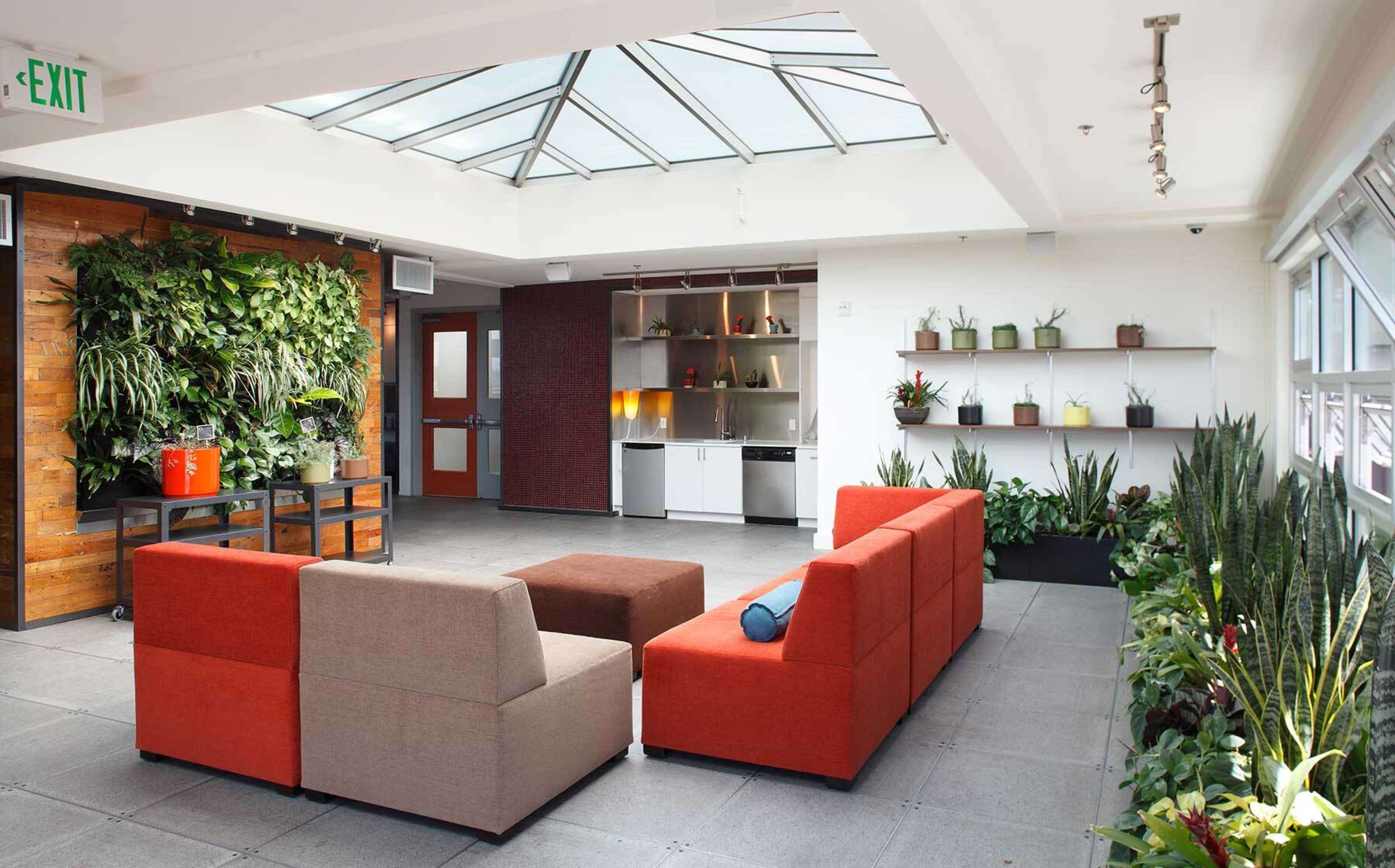 A bright indoor lounge area featuring a glass ceiling, modern furniture arranged in a seating configuration, and several plants displayed on shelves and walls.
