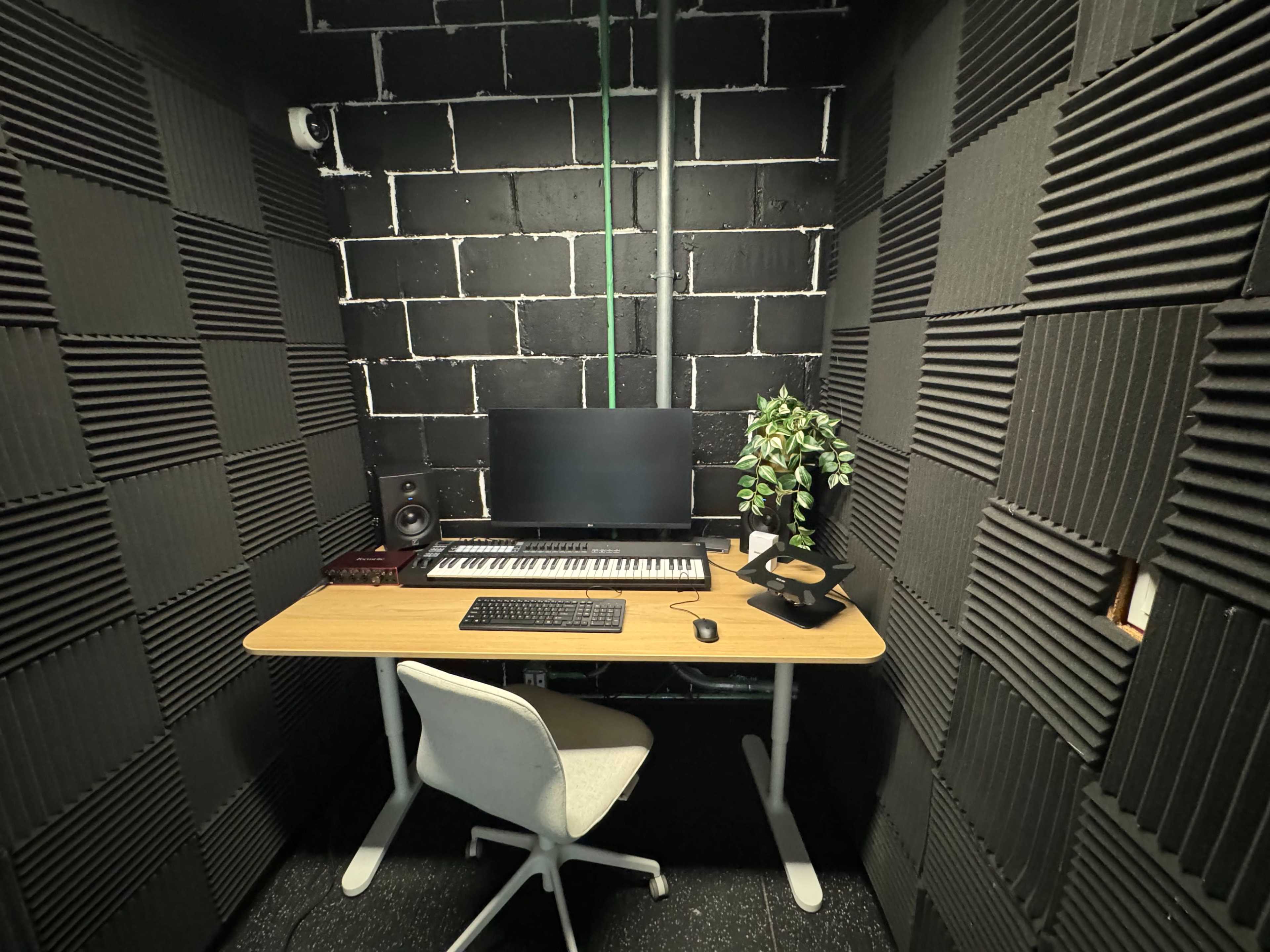 A small soundproofed studio features a desk with a computer monitor and keyboard, accompanied by a speaker and a potted plant against a black wall with sound-absorbing panels.